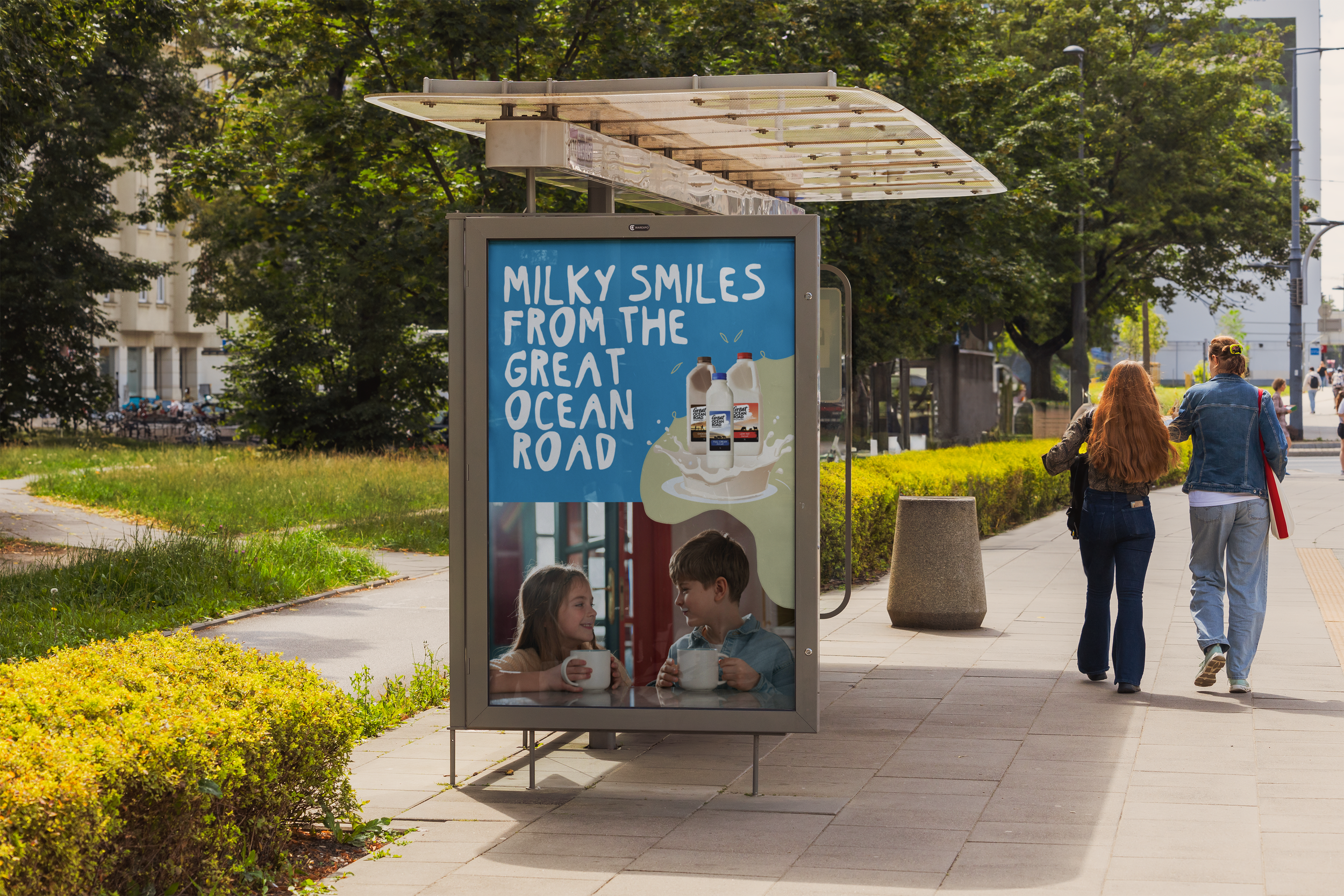 Public park with a vending machine advertizing milk, children sitting inside the machine drinking milk, two women walking along the sidewalk, trees and buildings in the background.
