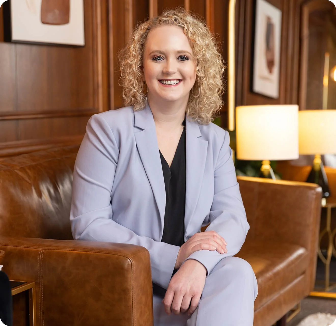 A woman with curly blonde hair sitting on a leather sofa in a warmly lit room, smiling at the camera, wearing a light gray blazer over a black top.