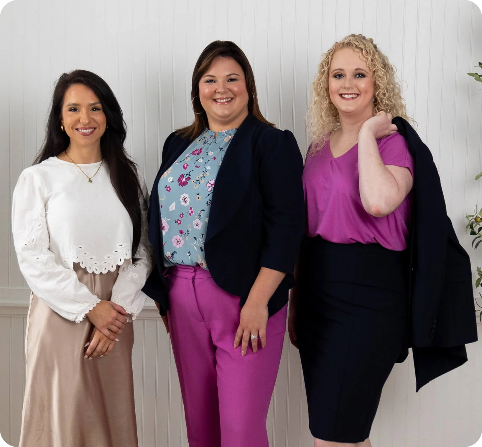 Three women standing in front of a white paneled wall, smiling, dressed in professional attire.