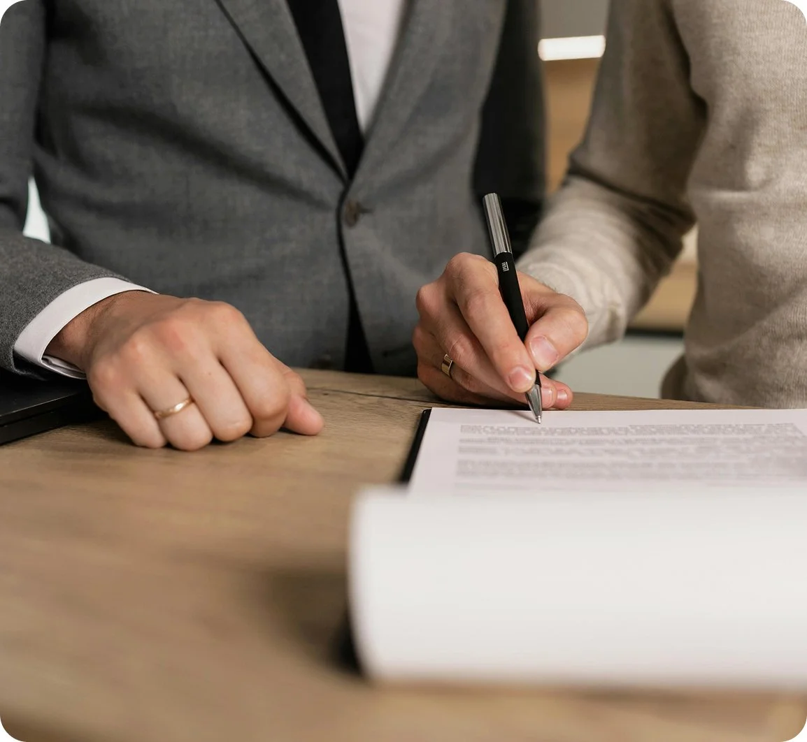 Two individuals are signing a document at a wooden table, with one person wearing a gray suit and the other wearing a beige sweater.