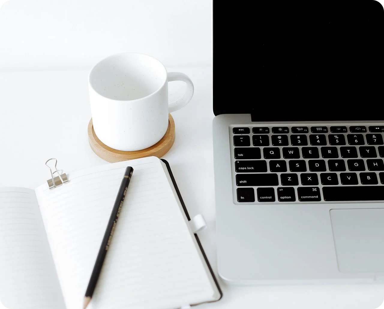 A workspace with a laptop, an open notebook with a pencil, a white coffee mug on a wooden coaster, and a binder clip on a white desk.