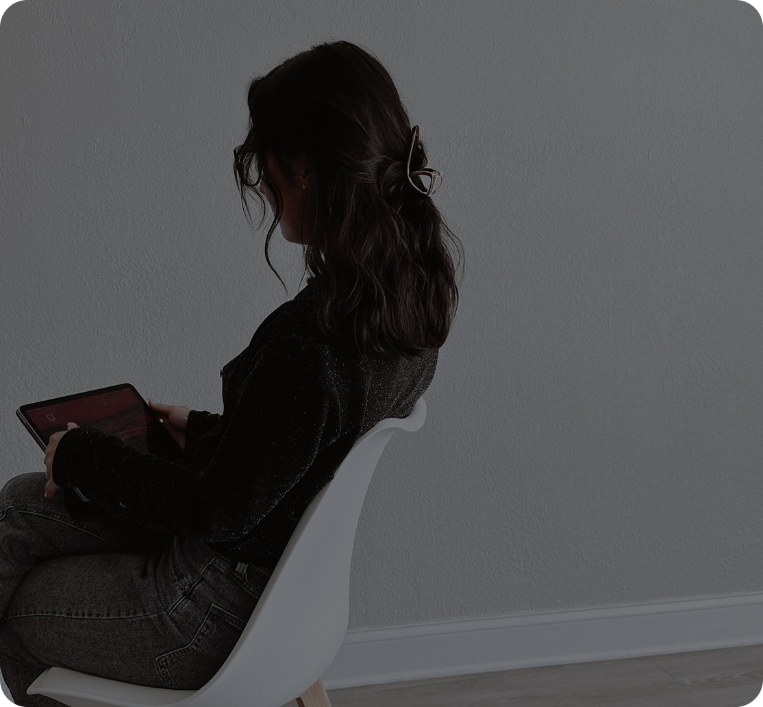 A woman with dark, wavy hair tied with a hair clip, sitting on a white chair and looking at her tablet against a plain wall background.