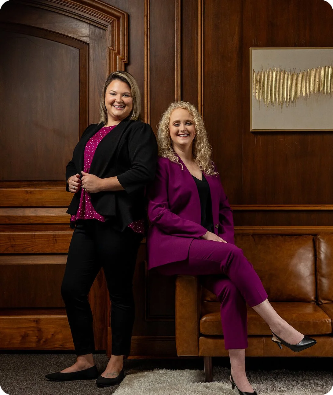 Two smiling women in business attire pose in an office with wooden paneling and a modern abstract painting on the wall.