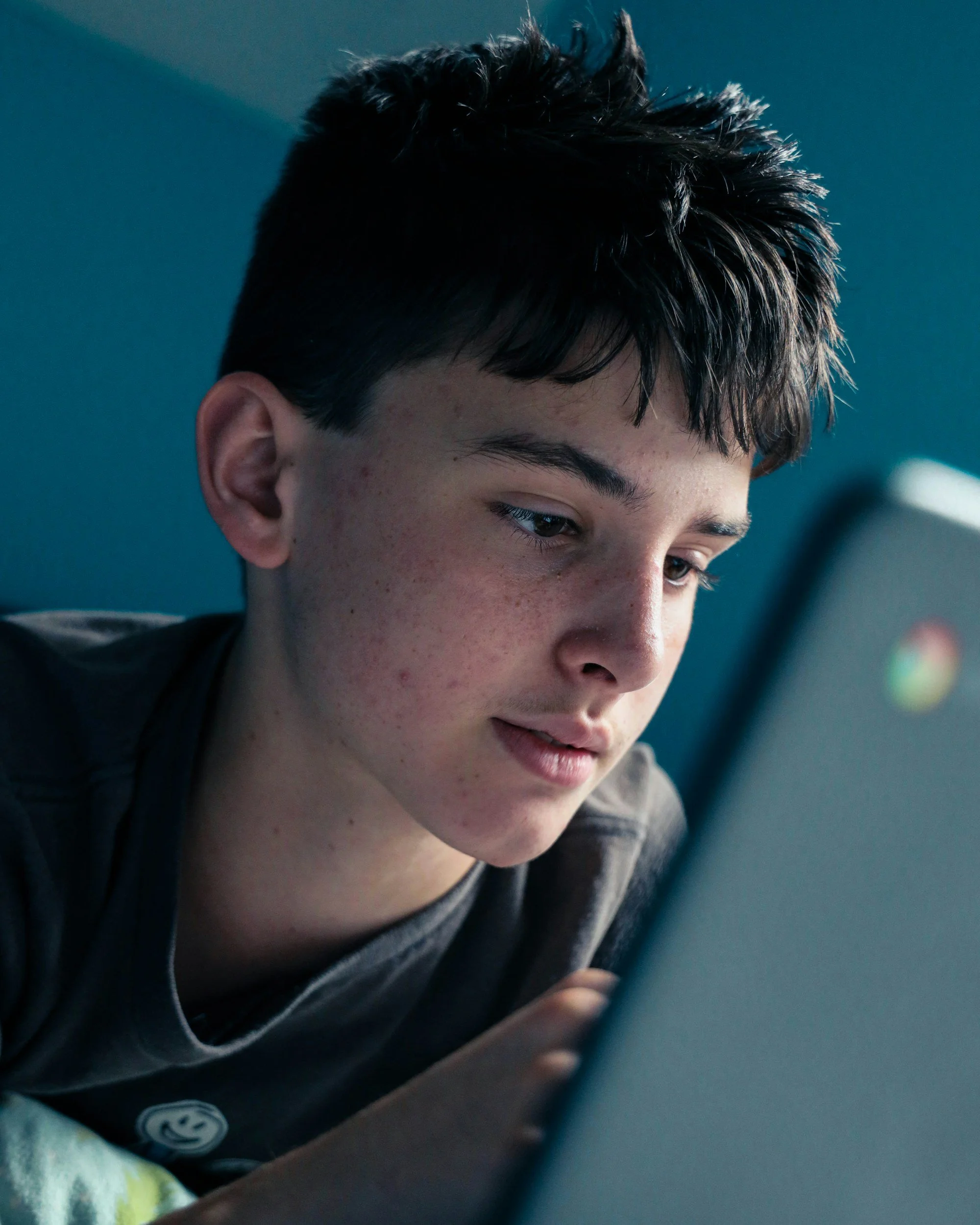 Close-up of a young person's face and hand, with short, dark, slightly messy hair, looking downward at a computer.