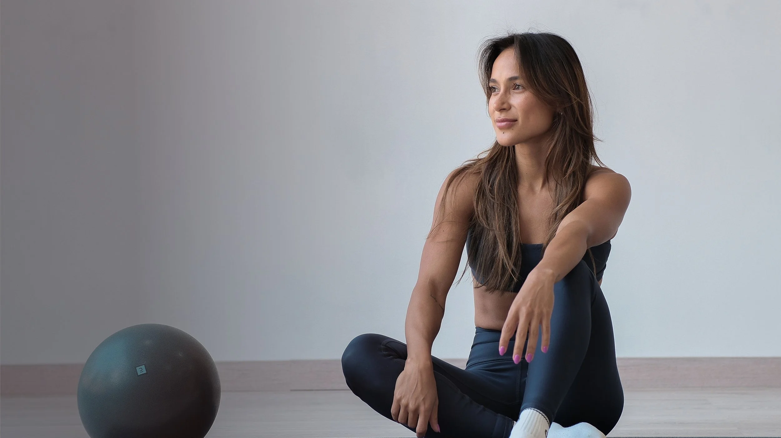 A woman sitting on the floor in a minimalistic room with light-colored walls, wearing athletic clothing, with a blue and black exercise ball nearby.