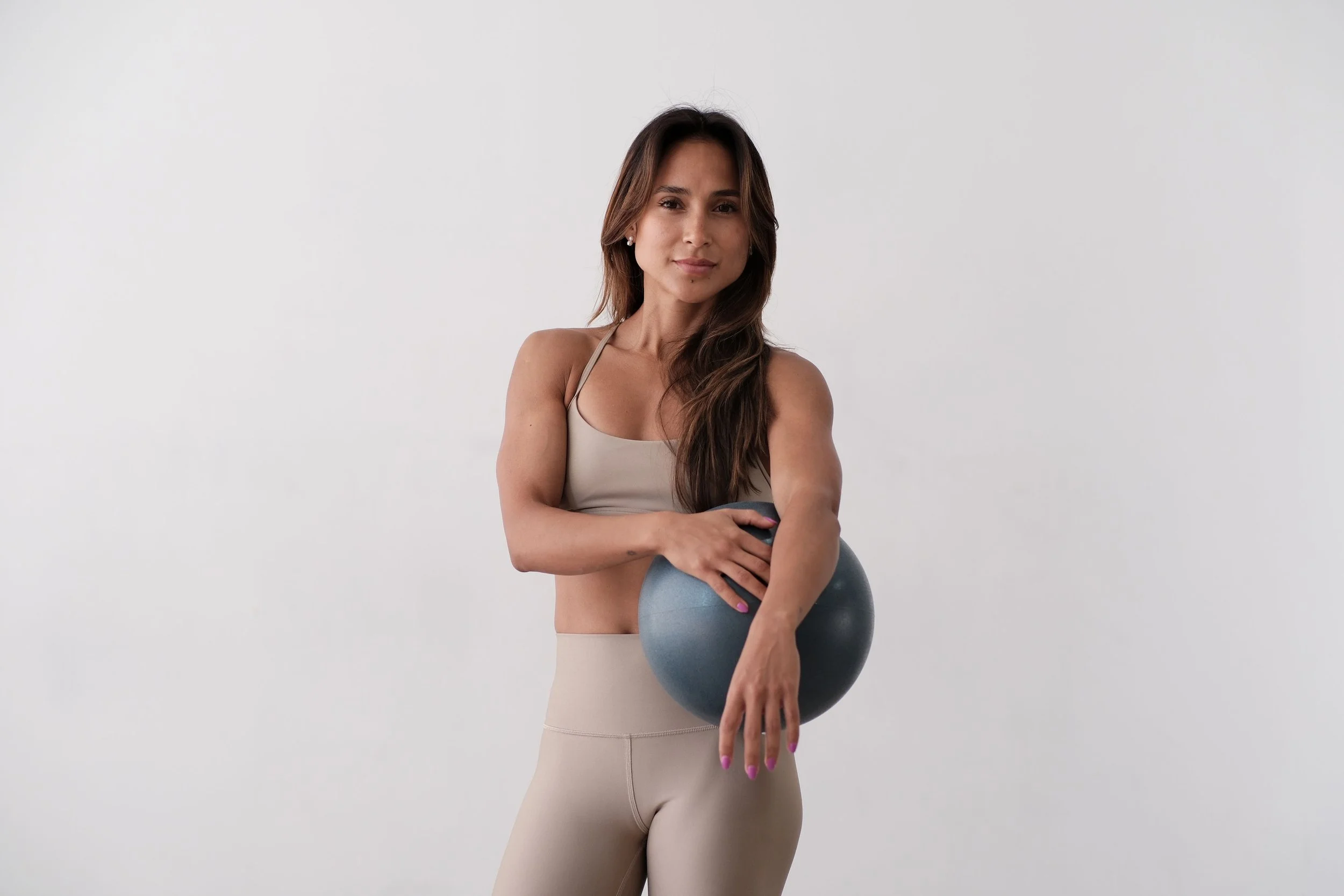 A woman in beige workout clothes holding a gray exercise ball against a plain white background.
