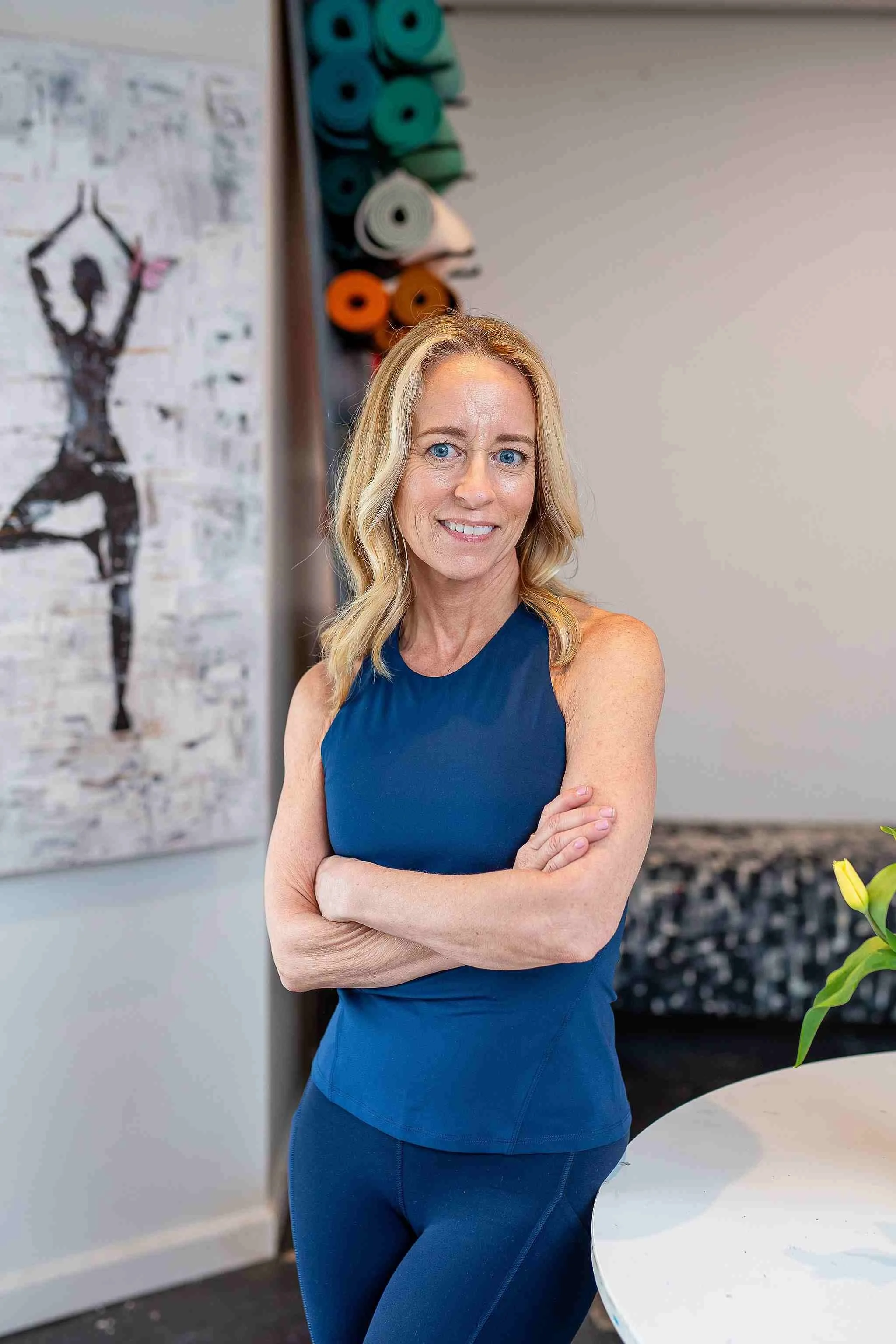 A woman with blonde hair and blue eyes wearing a blue sleeveless, standing with arms crossed and smiling in an indoor setting. In the background, there's a wall with colorful yoga mats lined up.