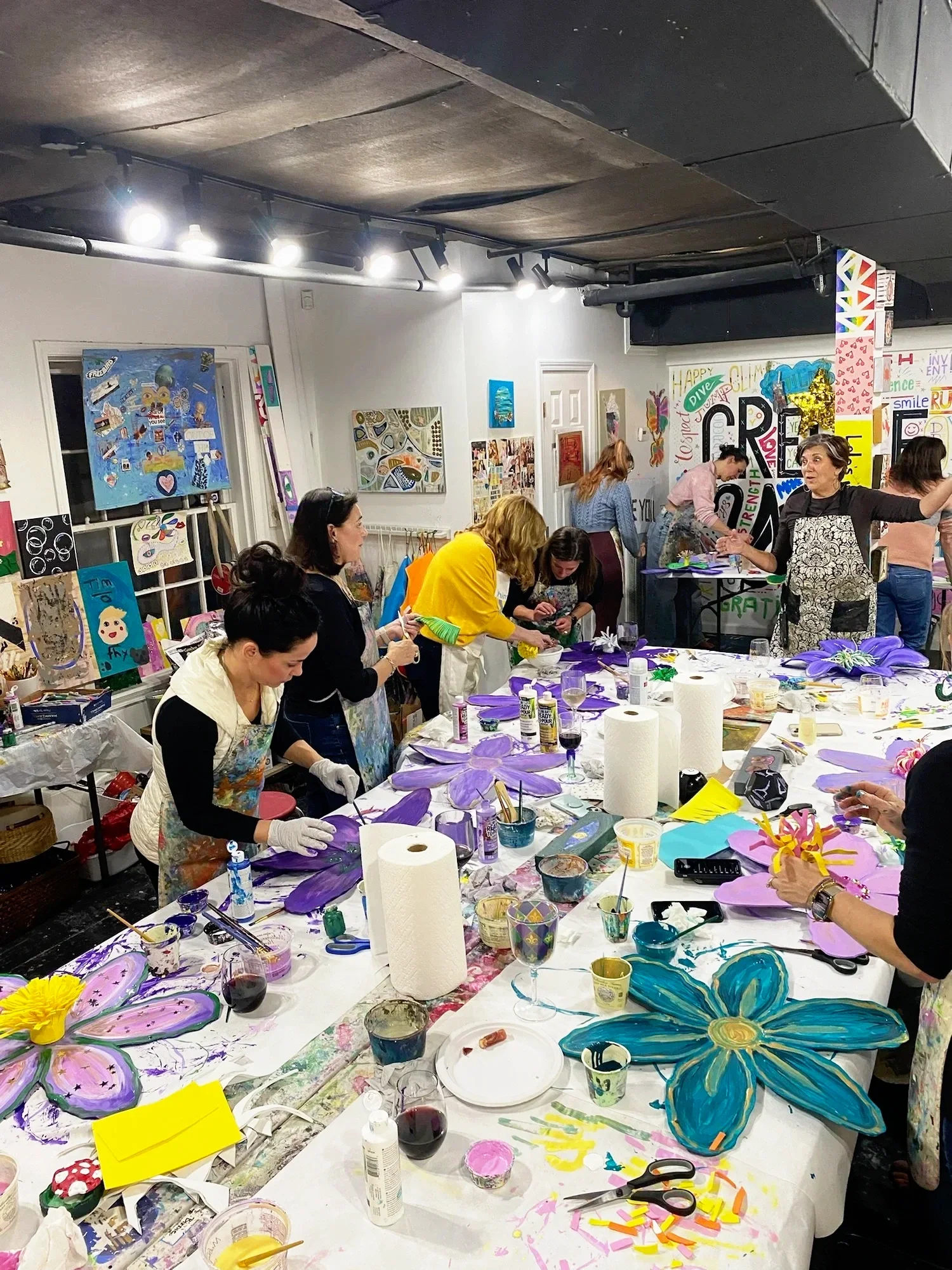 People participating in an art workshop, painting large colorful flowers. The room is decorated with vibrant artwork and filled with supplies such as paints and brushes.