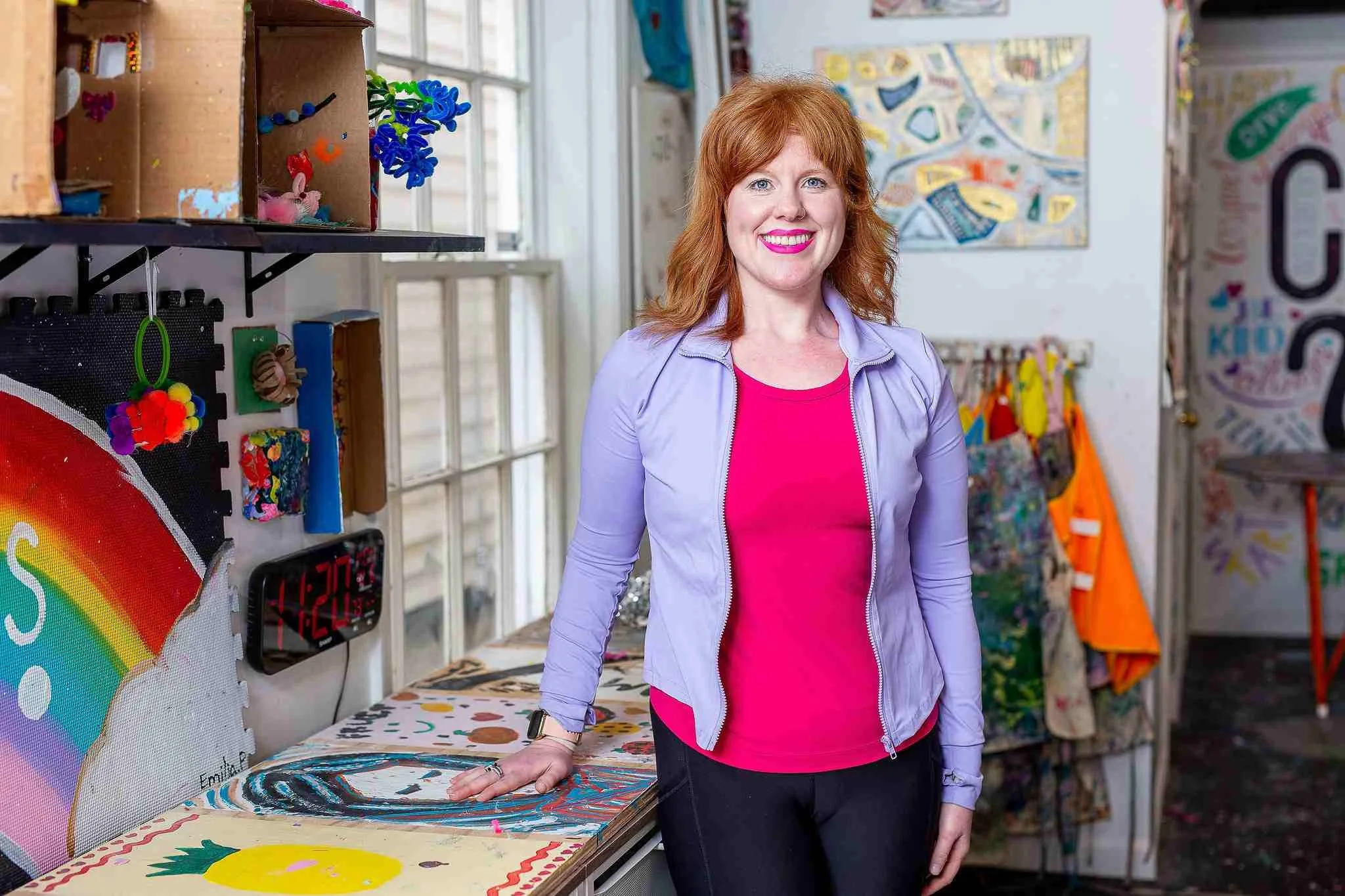 A woman with red hair and a bright smile standing in an art studio. She is wearing a purple jacket over a pink shirt, with her right hand resting on a painting on a table. Behind her are shelves with colorful art supplies and artworks on the walls.