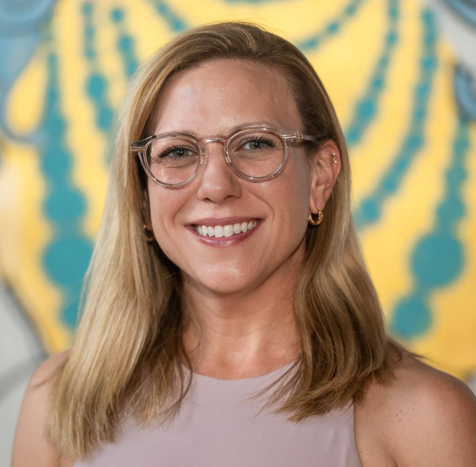 Portrait image of a woman with blonde hair, wearing glasses, smiling, earrings, and a sleeveless top, with a yellow and blue patterned background.