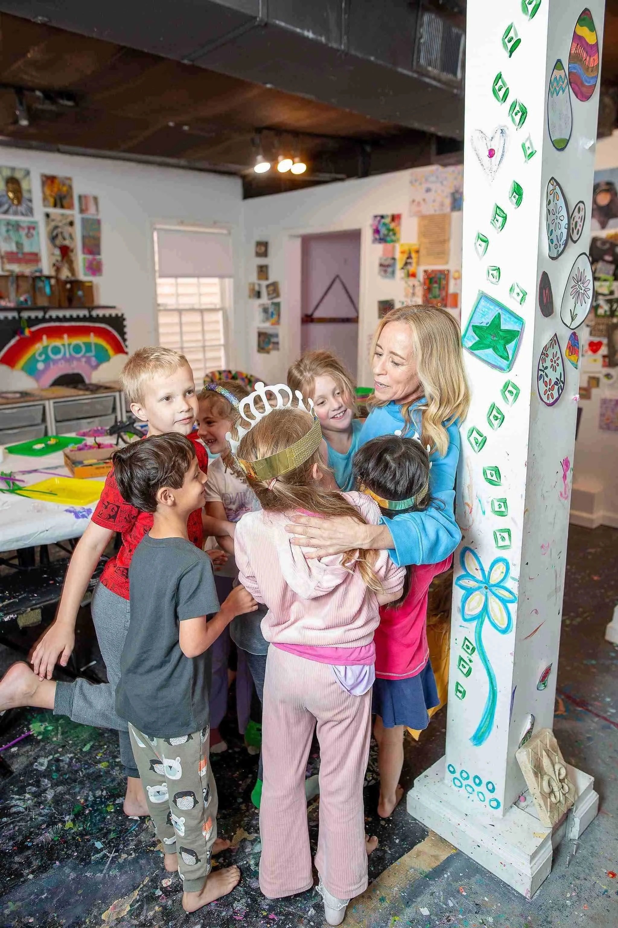 A group of children and a woman are gathered inside a colorful, art-filled room. The woman is hugging one of the children. The background showcases various artworks on the walls and a table with art supplies.