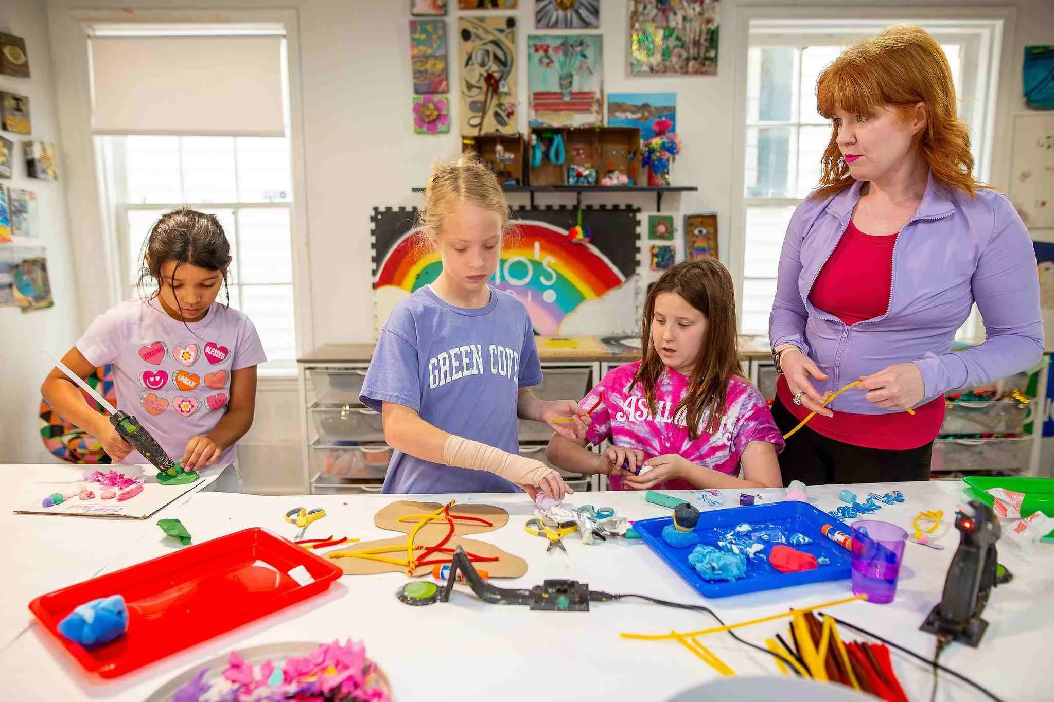 A group of young girls and a woman in a colorful arts and crafts classroom, working on creative projects with various craft supplies on the table.