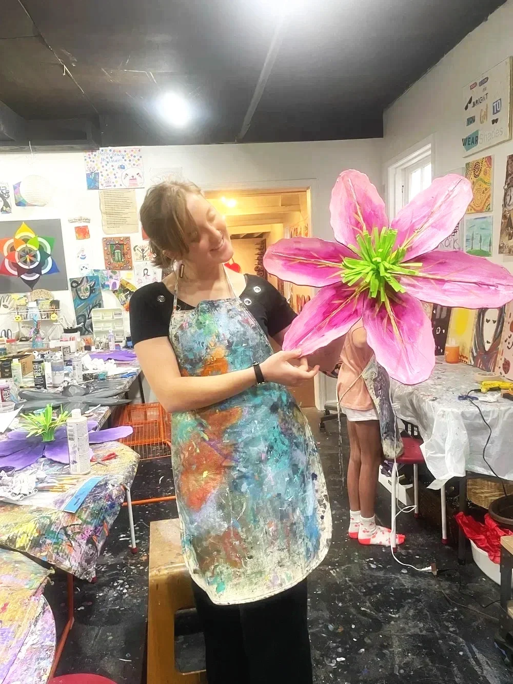 A woman in a splattered paint apron holding a large pink paper flower in an art studio, with colorful artwork and art supplies in the background.