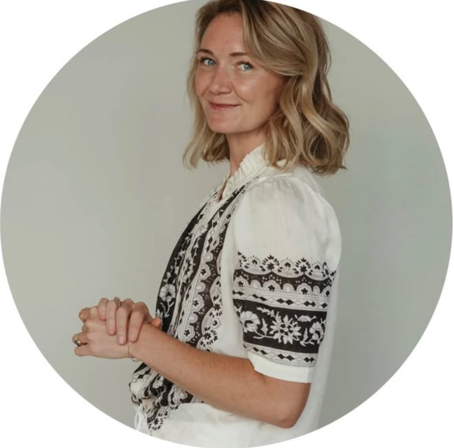 A woman with shoulder-length blonde hair, wearing a white blouse with black embroidered patterns, smiling and looking at the camera, standing against a light-colored background.