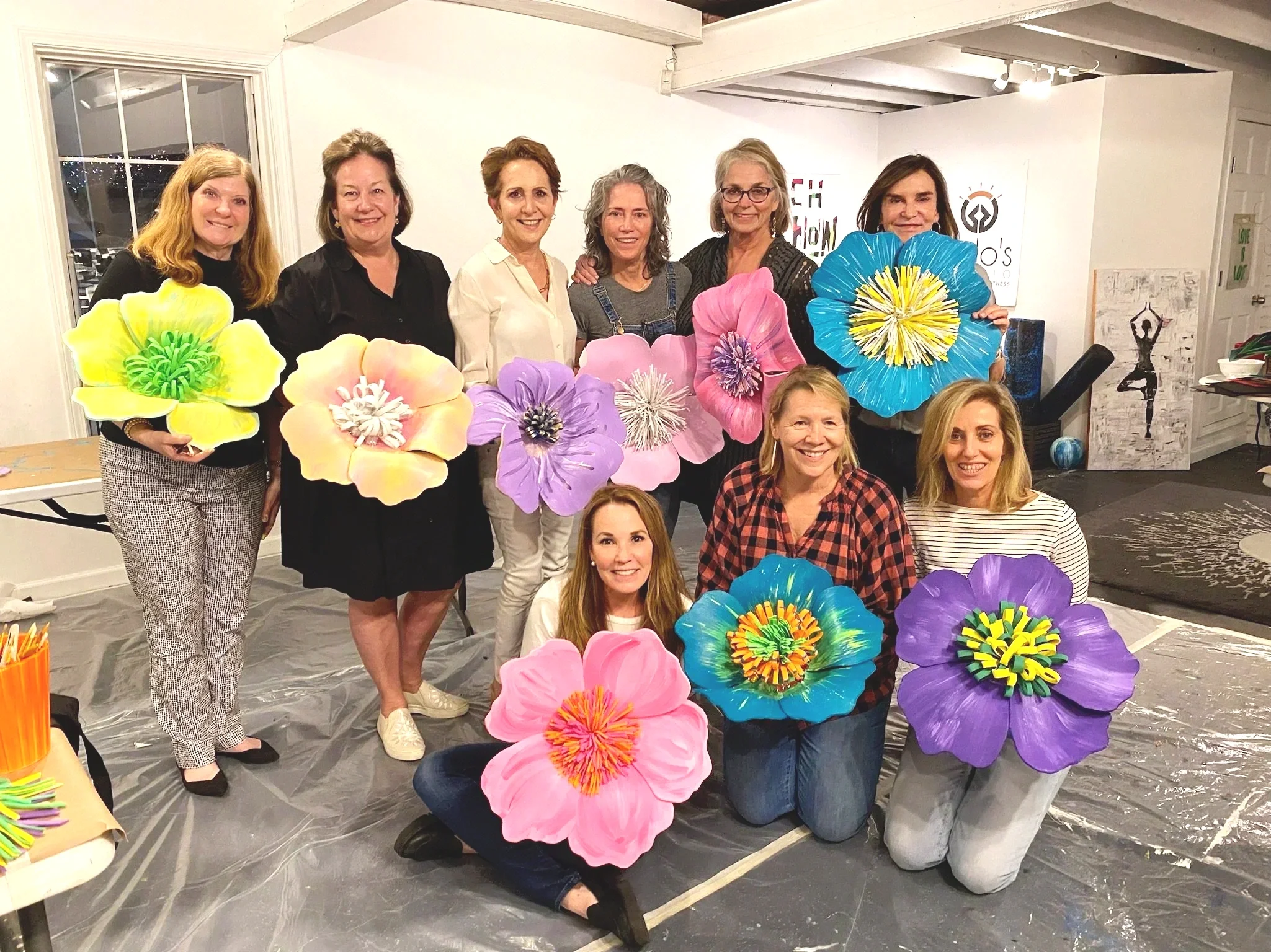 A group of women gathered together in an indoor space, holding large, colorful paper flowers. The women are smiling and are participating in a craft or art activity. The background shows a white wall with some artwork.