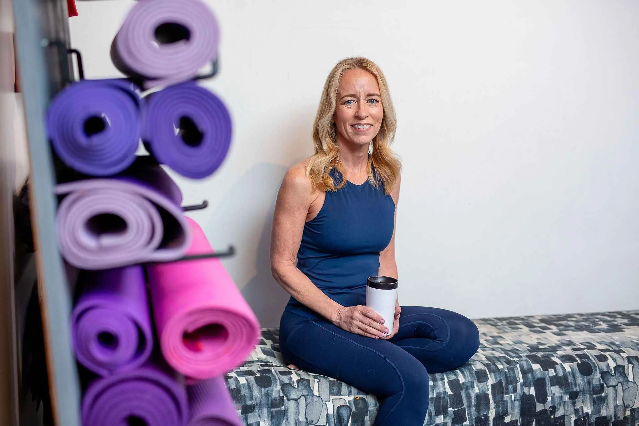 A woman is sitting on a patterned bed in a room, holding a white tumbler. There are purple and pink yoga mats rolled and stored on a rack nearby. The woman has blonde hair, blue eyes, and is wearing a sleeveless blue top and dark blue leggings.