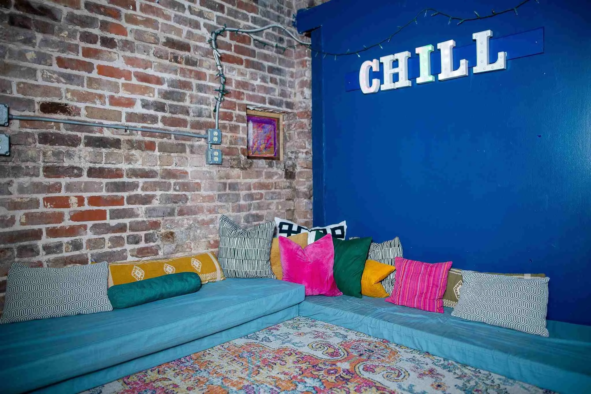 A cozy indoor lounge area with a vibrant blue wall decorated with a string of fairy lights and a large illuminated sign spelling 'CHILL.' There is a long, turquoise sofa adorned with multiple colorful and patterned pillows, placed on a multicolored ornate rug. The background features a brick wall