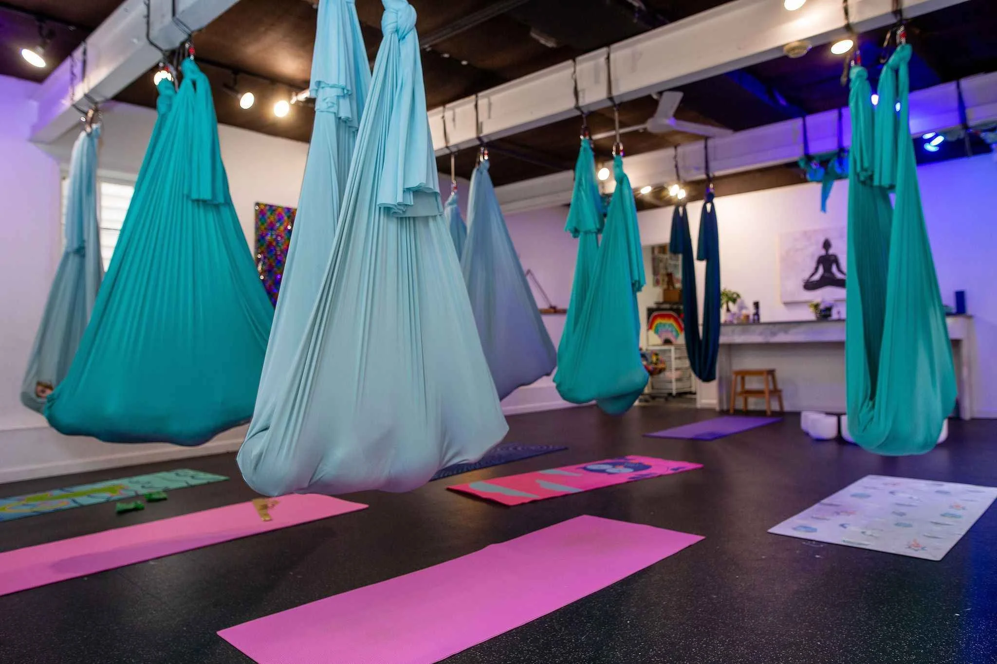 Indoor yoga studio with multiple hanging aerial silk hammocks in shades of blue and teal, yoga mats and colorful artwork on the walls, and soft lighting creating a peaceful atmosphere.