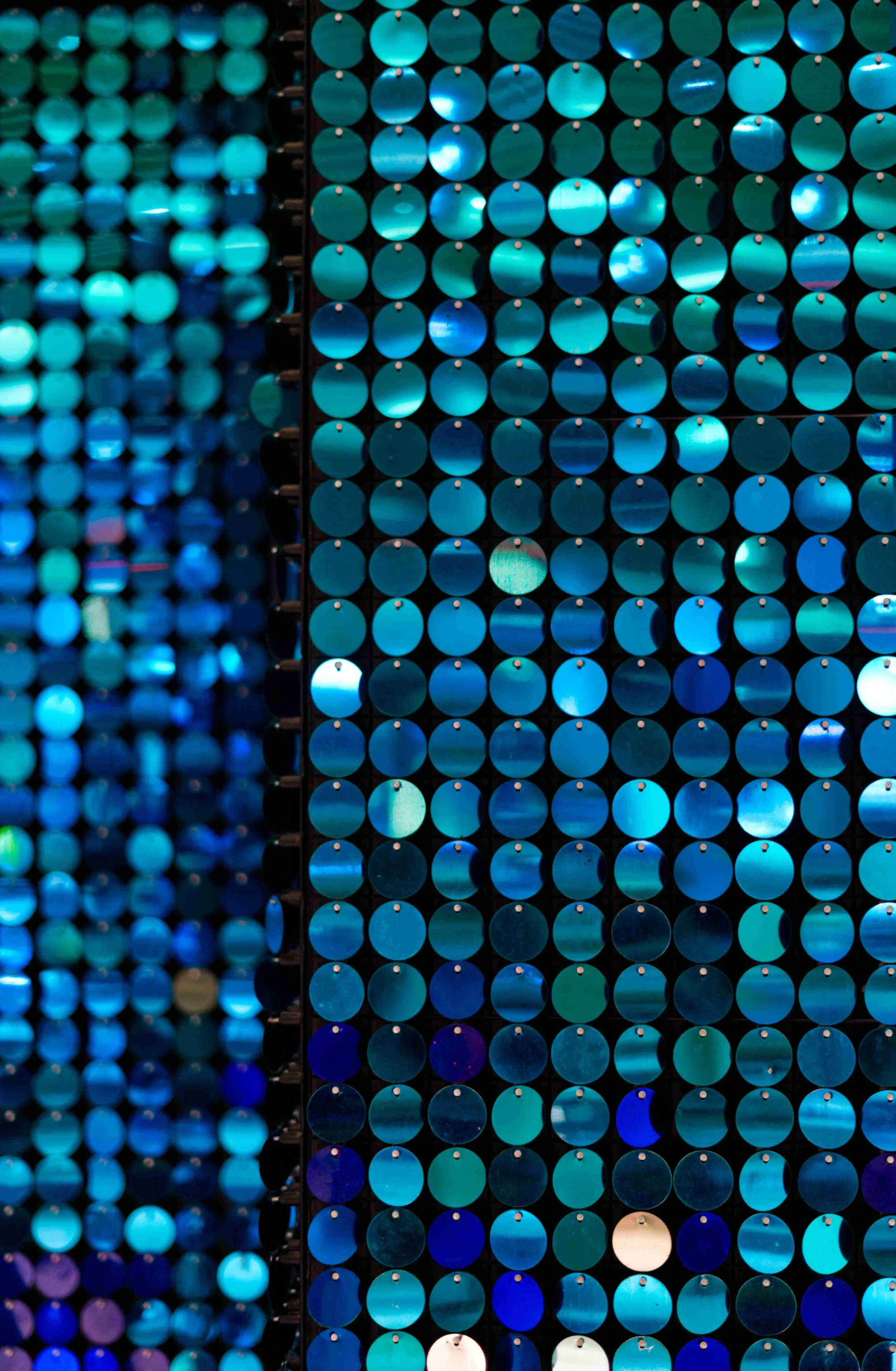 Close-up of a wall display made of numerous round, blue, and teal reflective discs arranged neatly in rows, with some discs reflecting lights and colors.