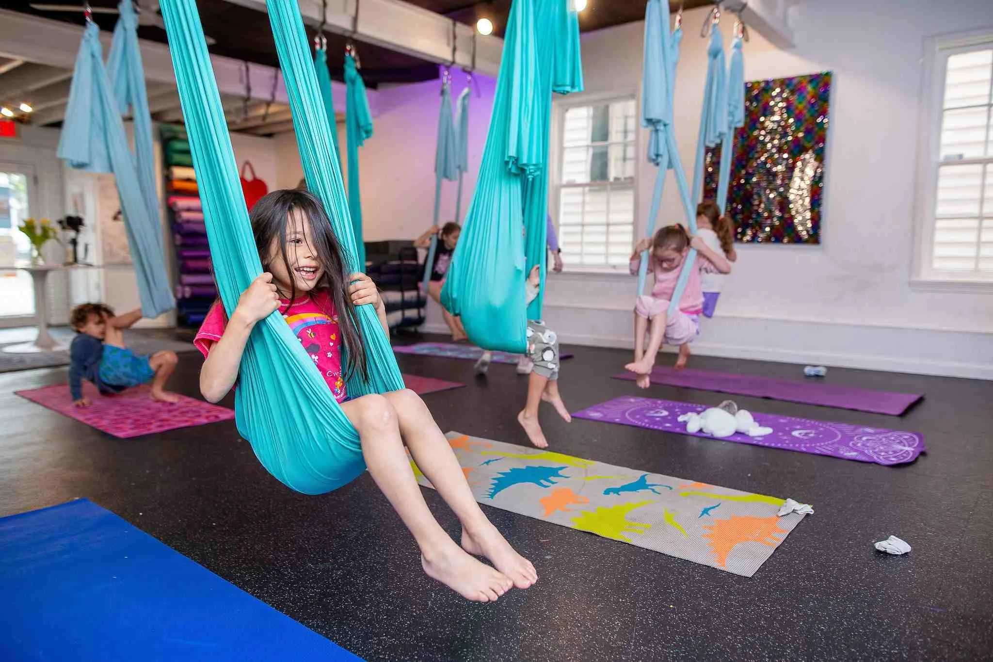 Children engaging in aerial yoga using blue fabric swings in a bright studio with yoga mats and colorful wall art.