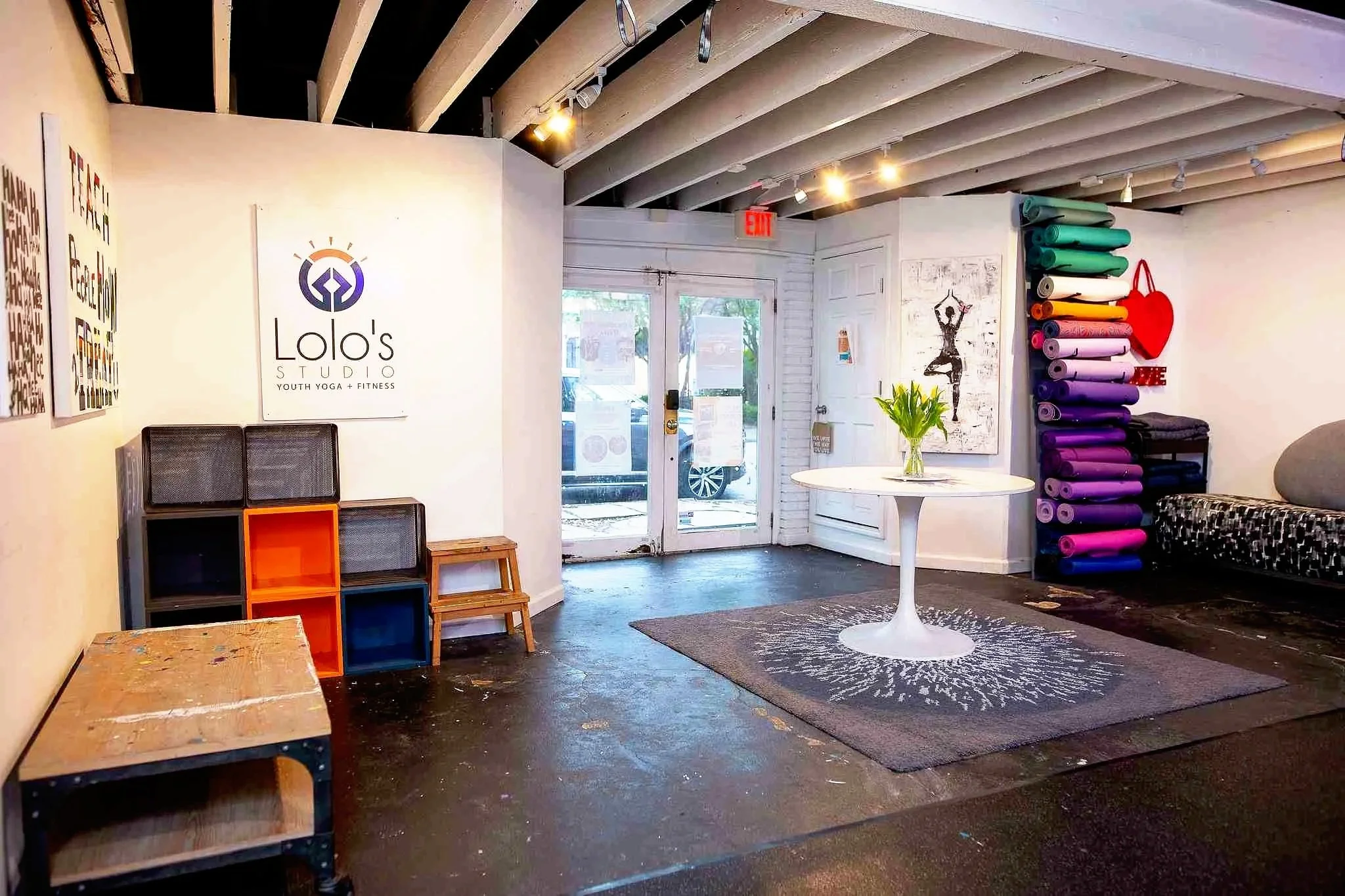 Interior of a fitness studio with yoga mats stacked on the wall, a small round table with a plant, artwork on the walls, and a door with windows leading outside.