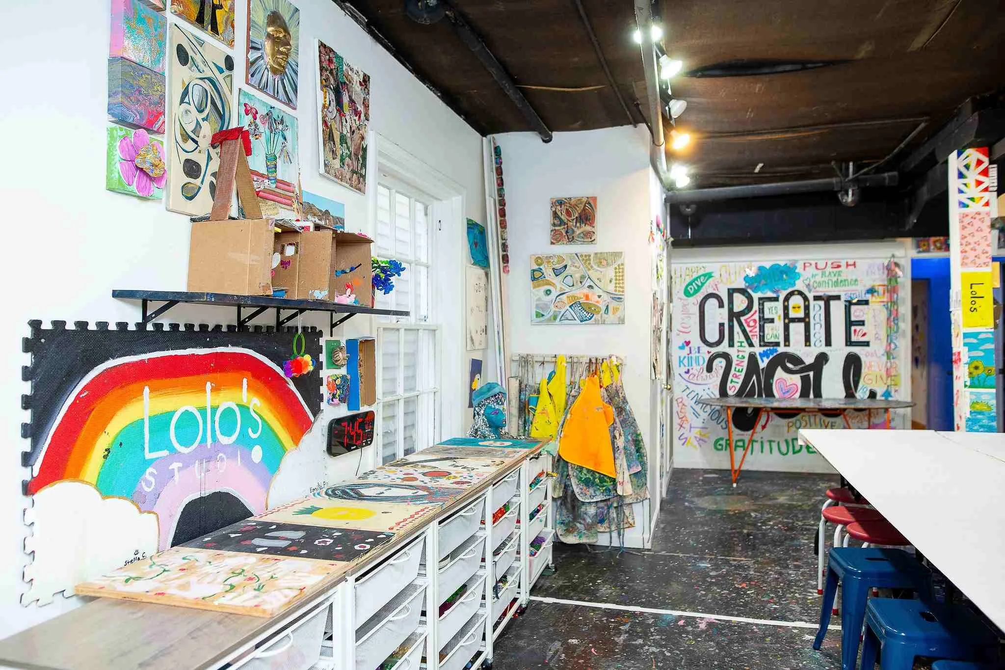 An art studio filled with colorful paintings, drawings, and crafts on tables and walls. There's a rainbow-themed sign on the wall. Various art supplies and decorative items are scattered throughout the room.