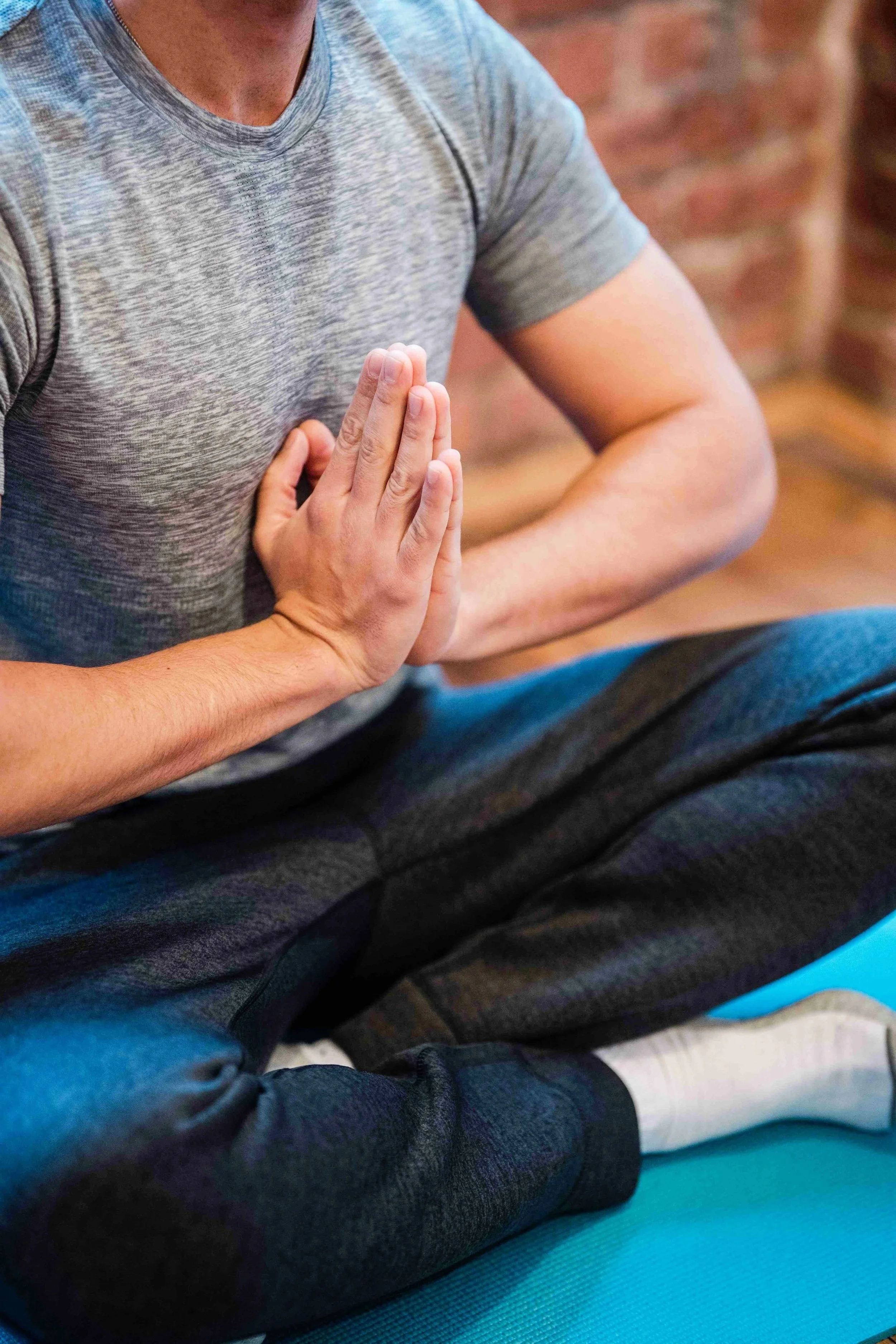 A person wearing a gray t-shirt is seated on a blue exercise mat with their hands pressed together in a prayer position on their chest, practicing yoga or meditation.