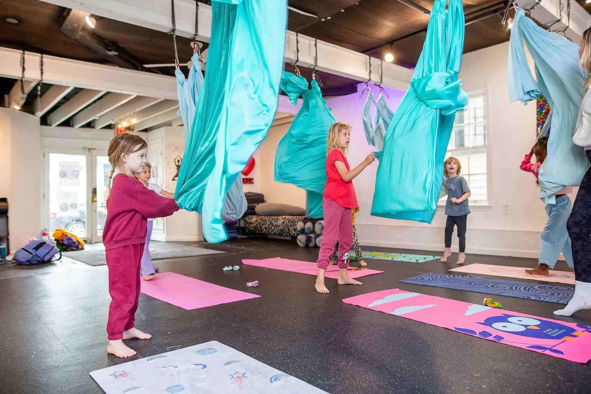 Children participating in a yoga class with yoga mats and aerial hammocks hanging from the ceiling in a room with large windows and wooden ceiling.