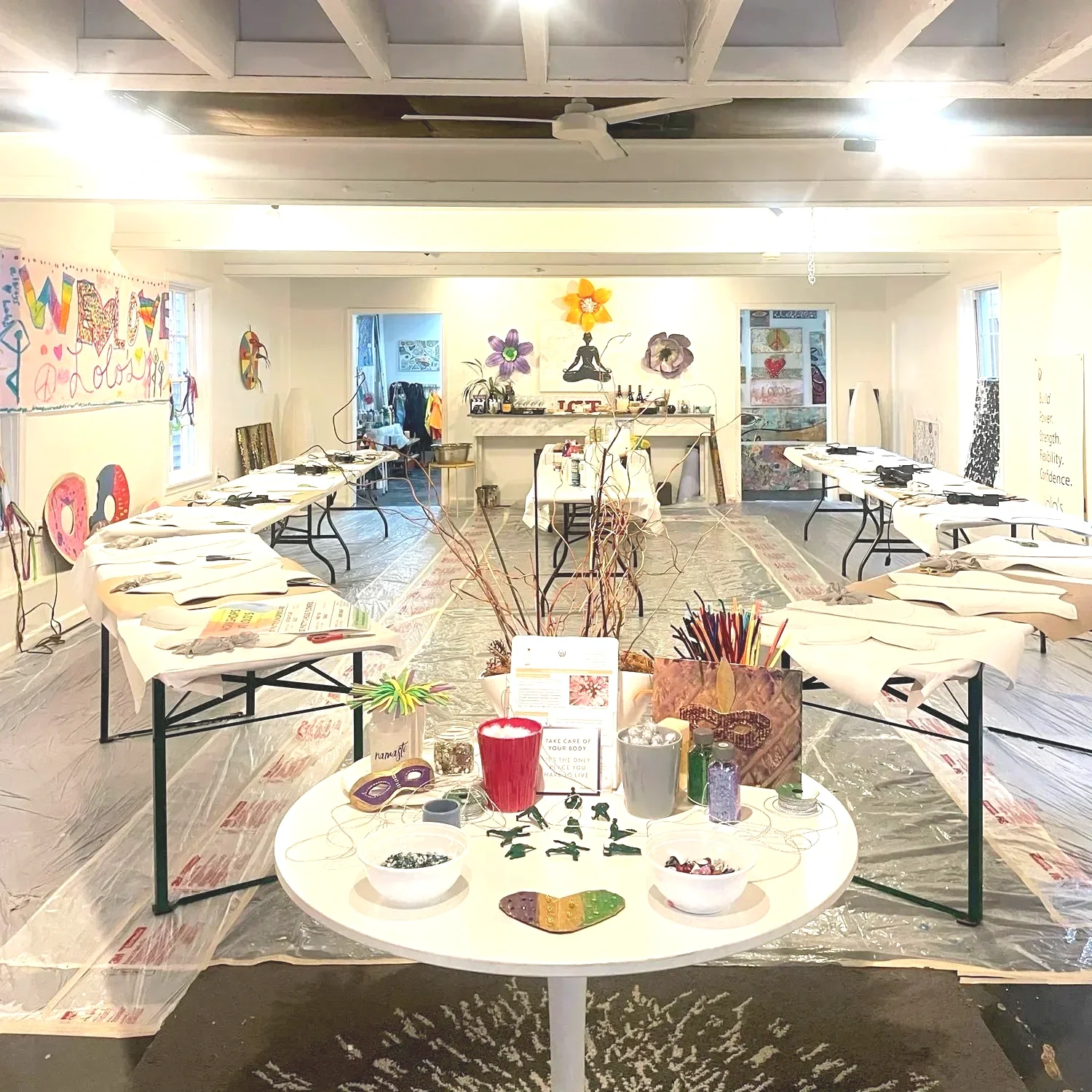 Indoor art studio or workshop space with tables covered in paper and art supplies, colorful wall art, and various decorations.