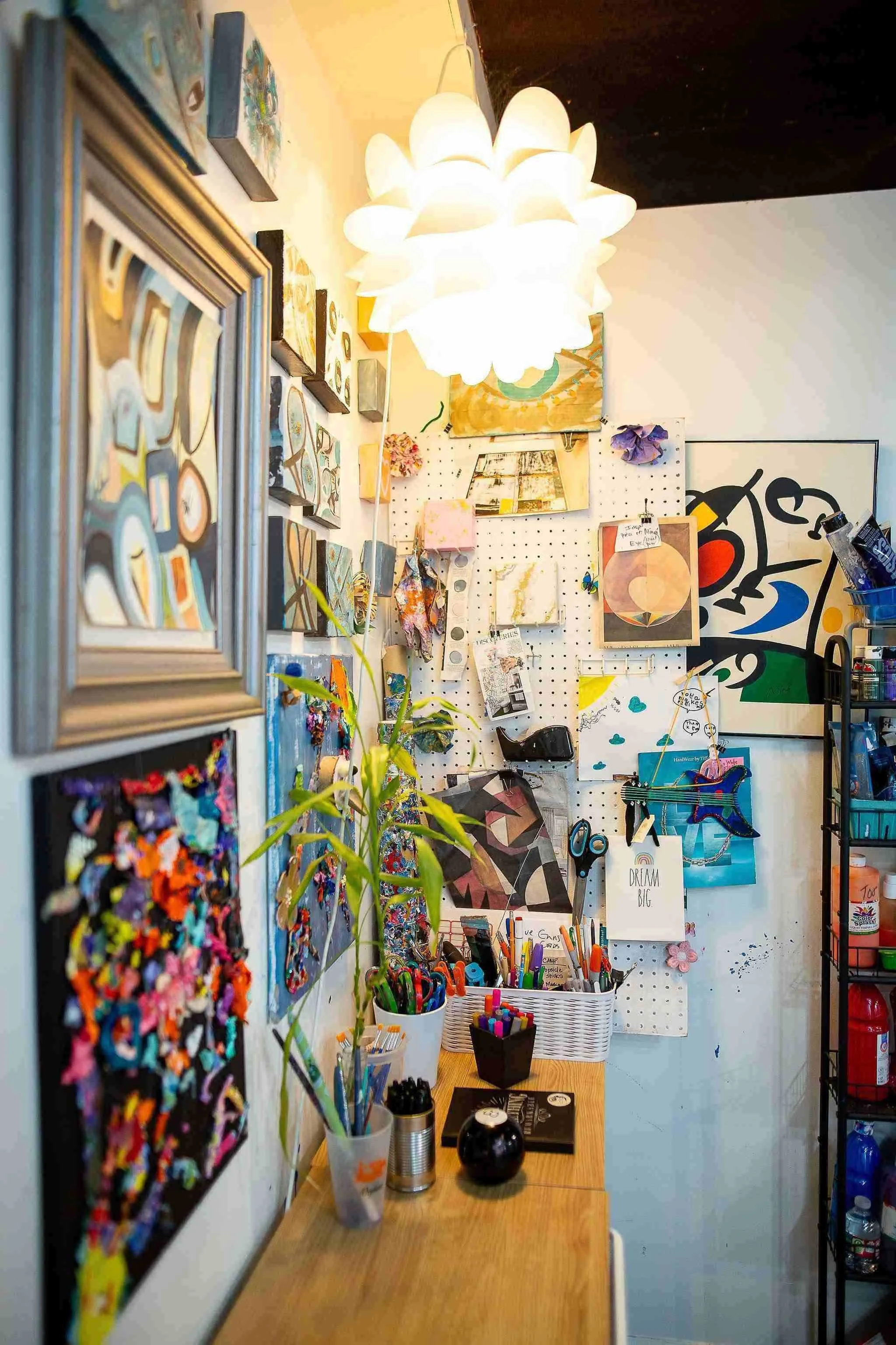 A craft or art studio workspace with a wooden table, various art supplies including markers, scissors, and paint, decorated artwork on the wall, and a hanging ceiling light.
