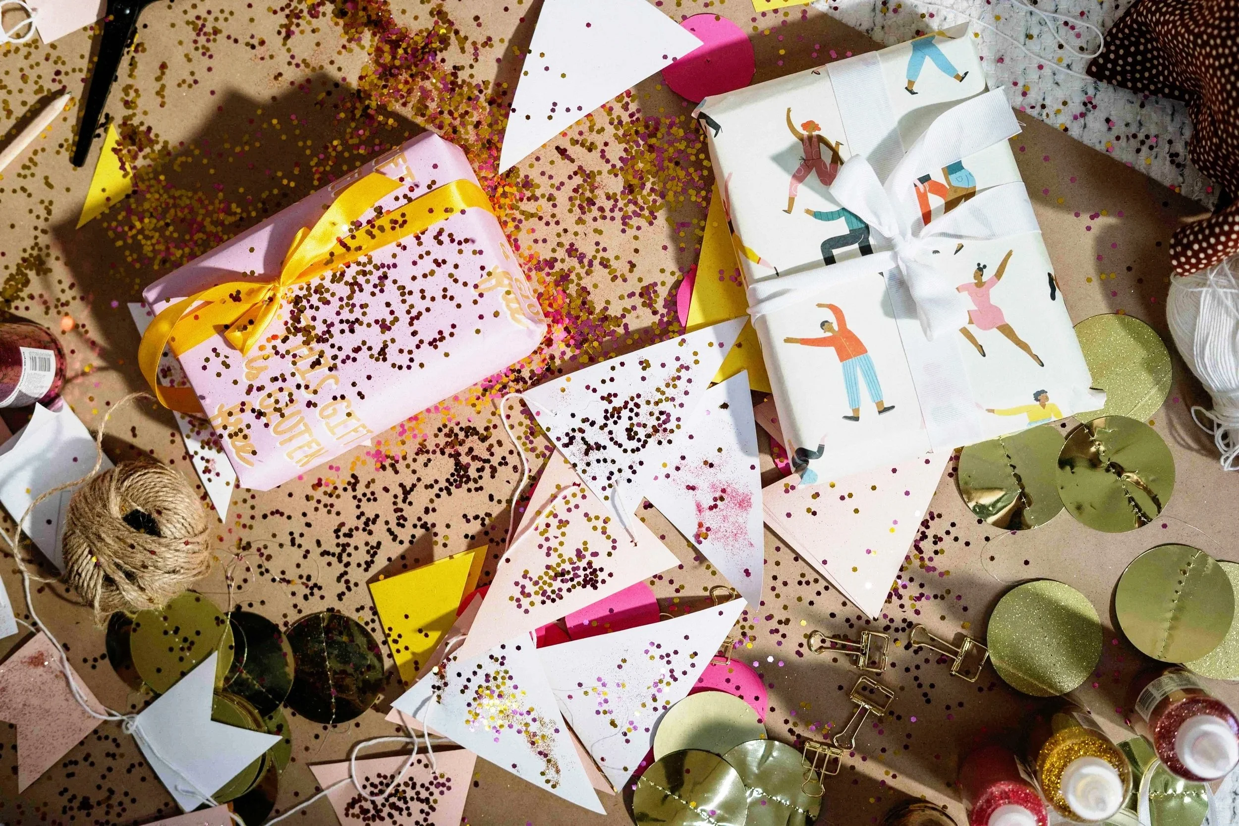A cluttered table with wrapped gifts, confetti, gold decorations, twine spool, and various craft supplies and tools scattered around.