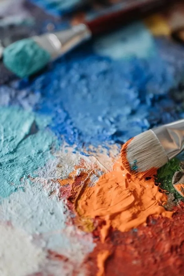 Close-up of a painter's palette with mixed blue, orange, and green oil paints, and a paintbrush resting on the palette.