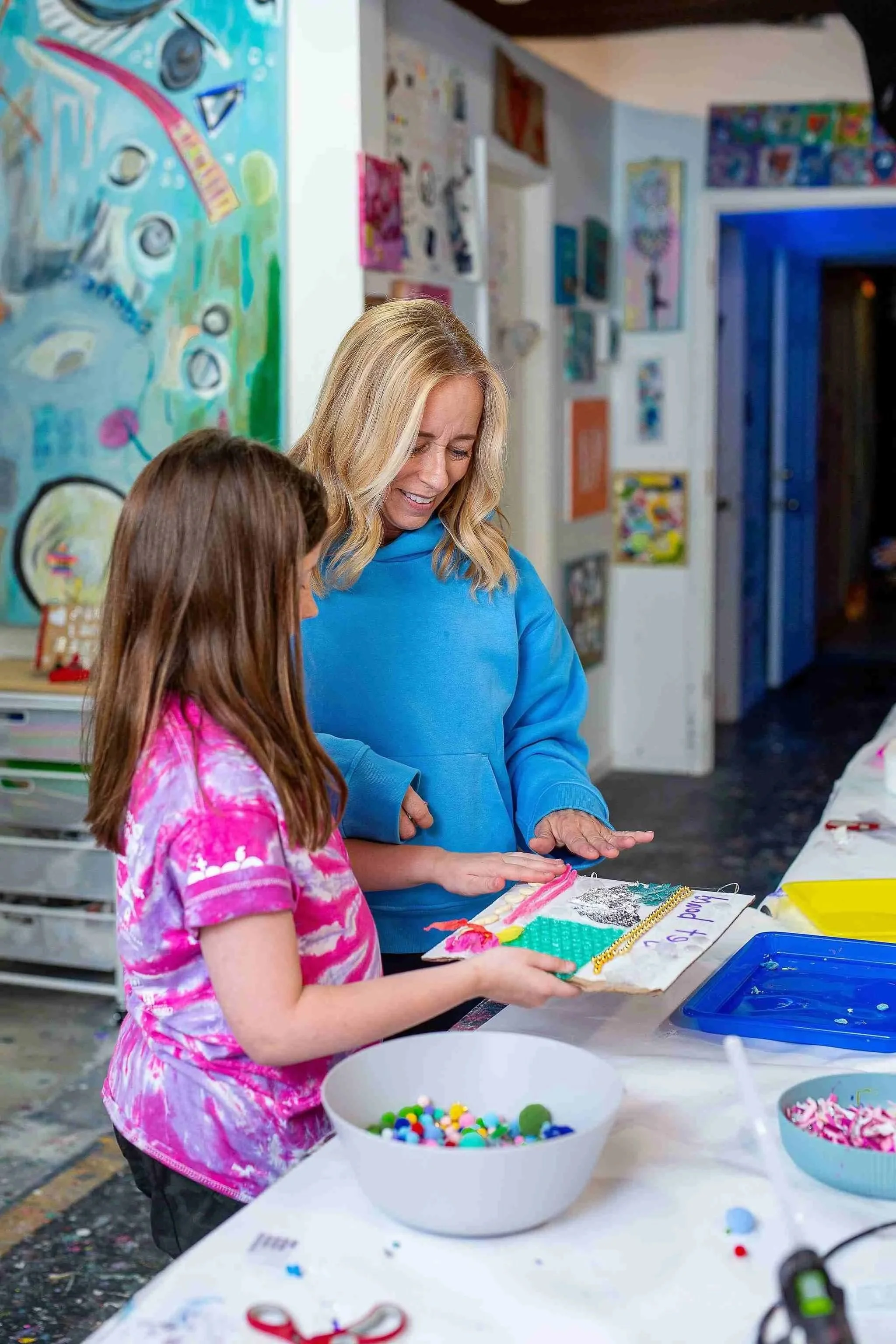 A woman and a girl are making a craft project together at a table, with bowls of colorful beads and craft supplies in front of them.