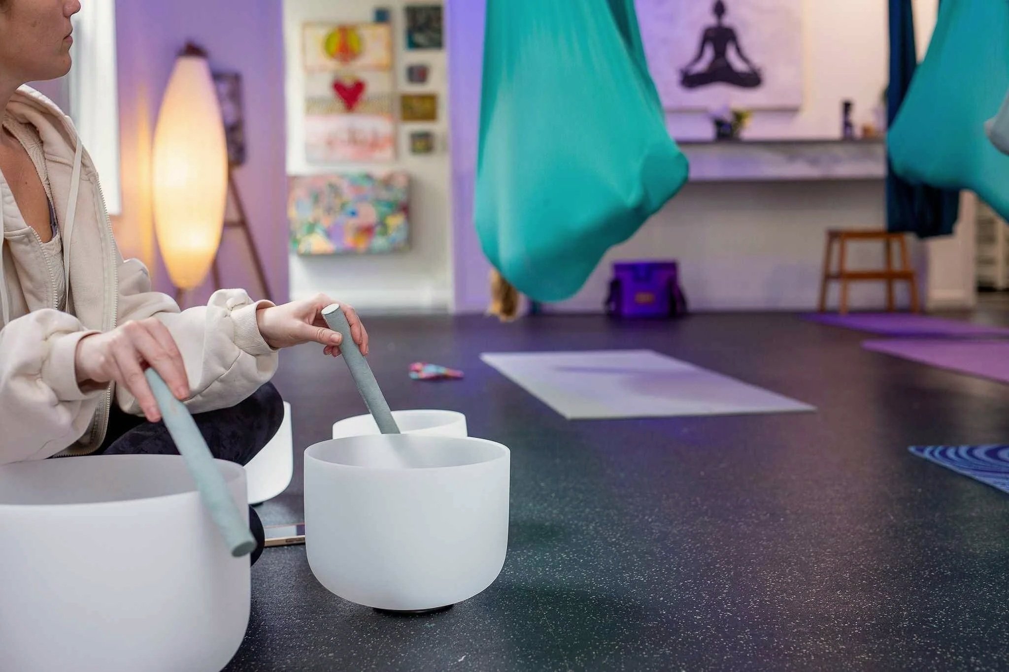 Close-up of a person in a beige hoodie playing singing bowls in a yoga studio with yoga mats on the floor.