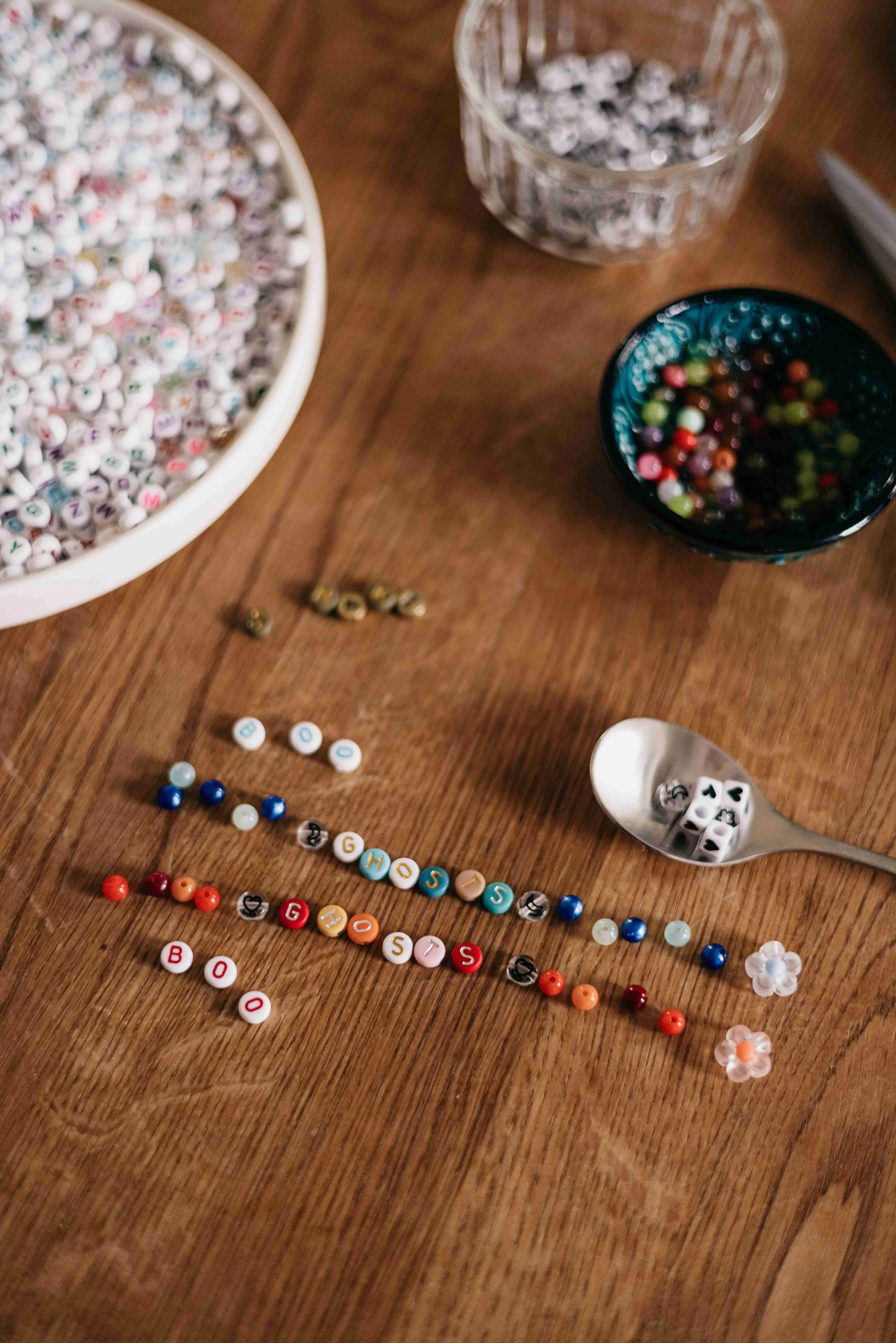 A wooden table with scattered colorful letter beads, white dice, and small jars filled with decorative beads and small objects.