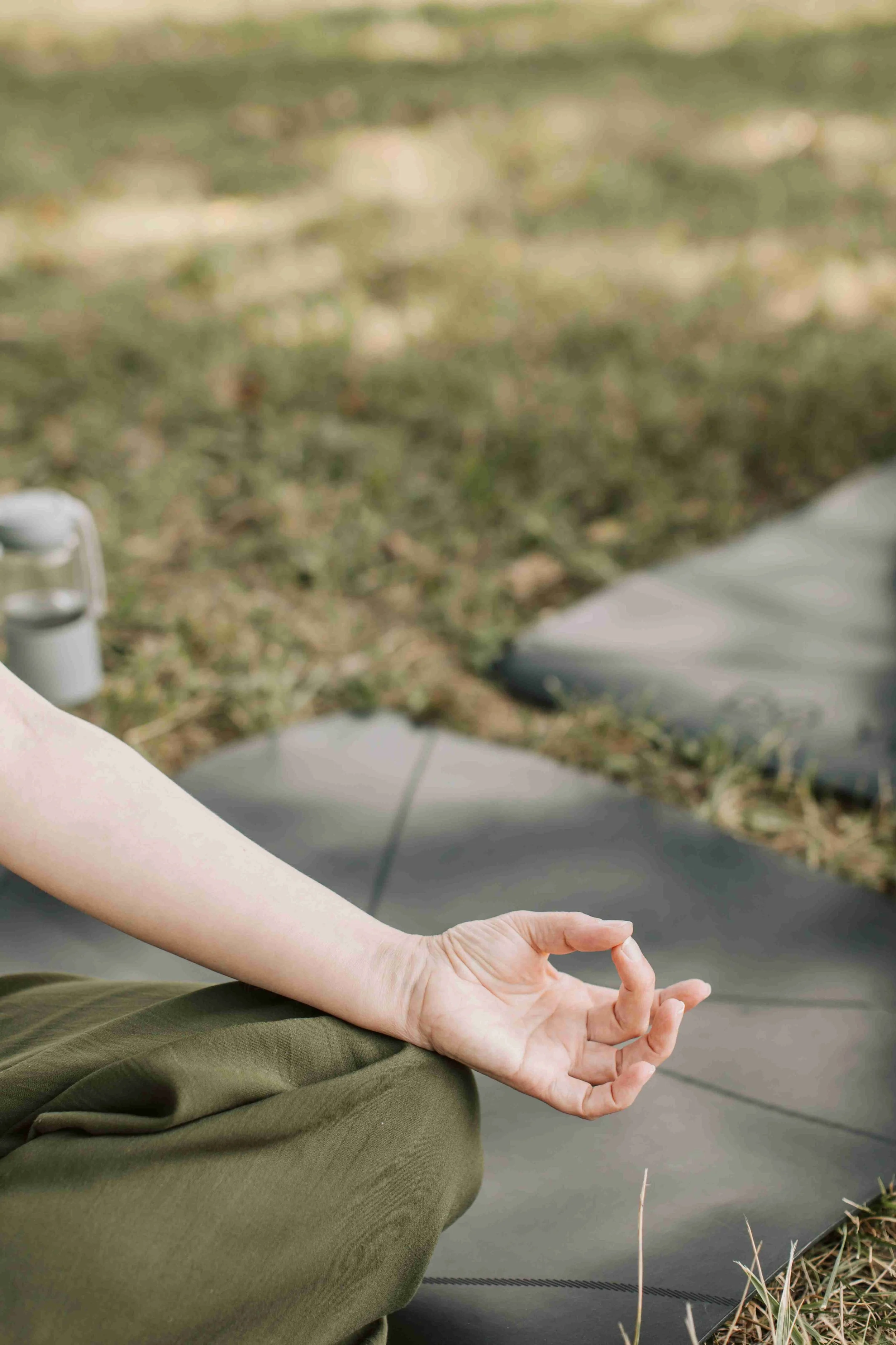 A person practicing meditation outdoors, seated on a yoga mat with their hand in a mudra position, surrounded by grass and natural landscape.