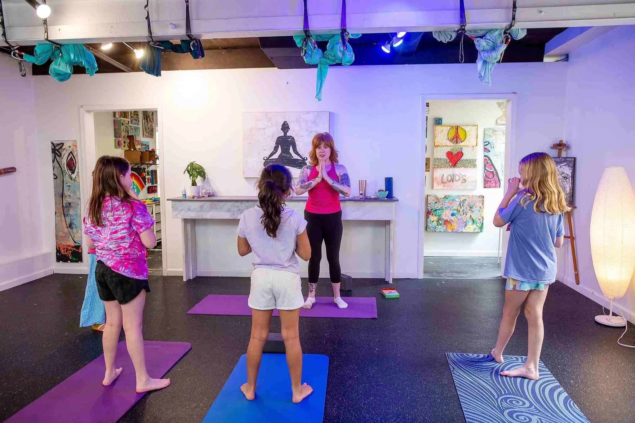 A woman leading a yoga class for children in a bright, art-filled room. The instructor is standing on a purple yoga mat, with six children standing on colored yoga mats in front of her, each in a yoga pose. Artworks adorn the walls, and fabric swings hang from the ceiling.