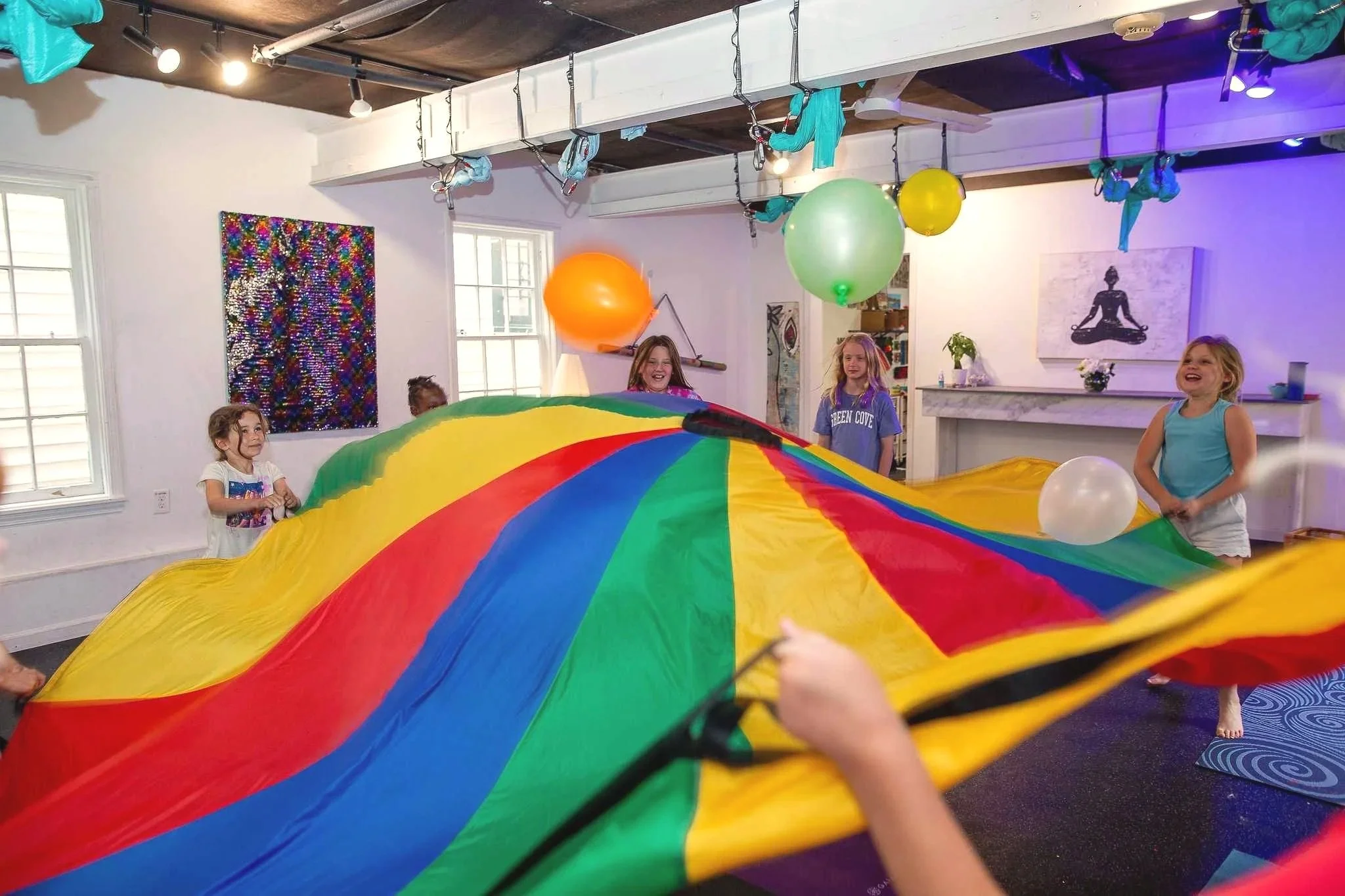 Children playing with a multicolored parachute indoors, with balloons and artwork visible in the background.