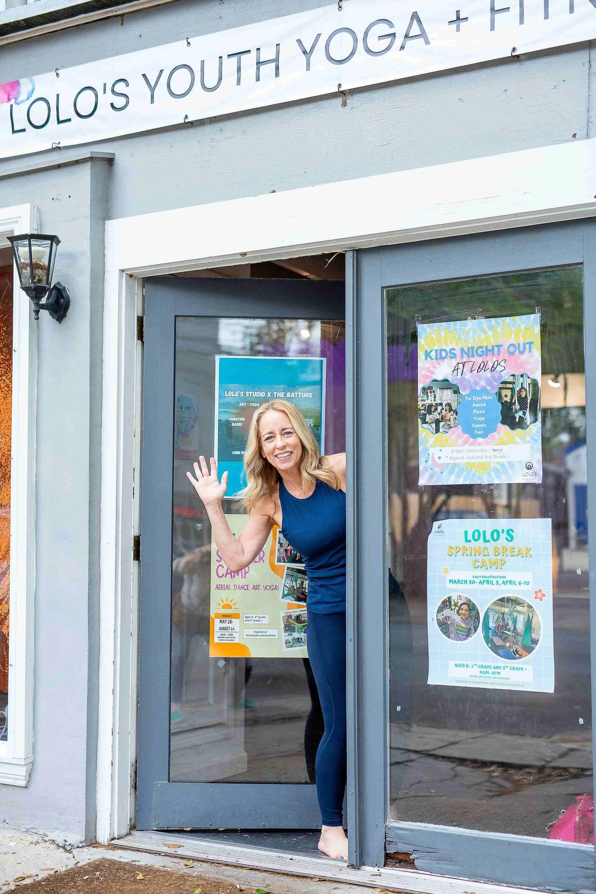 A woman smiling and waving at the entrance of a building with promotional posters for kids' events and camps, including a kids' night out and spring break camp at Lolo's.