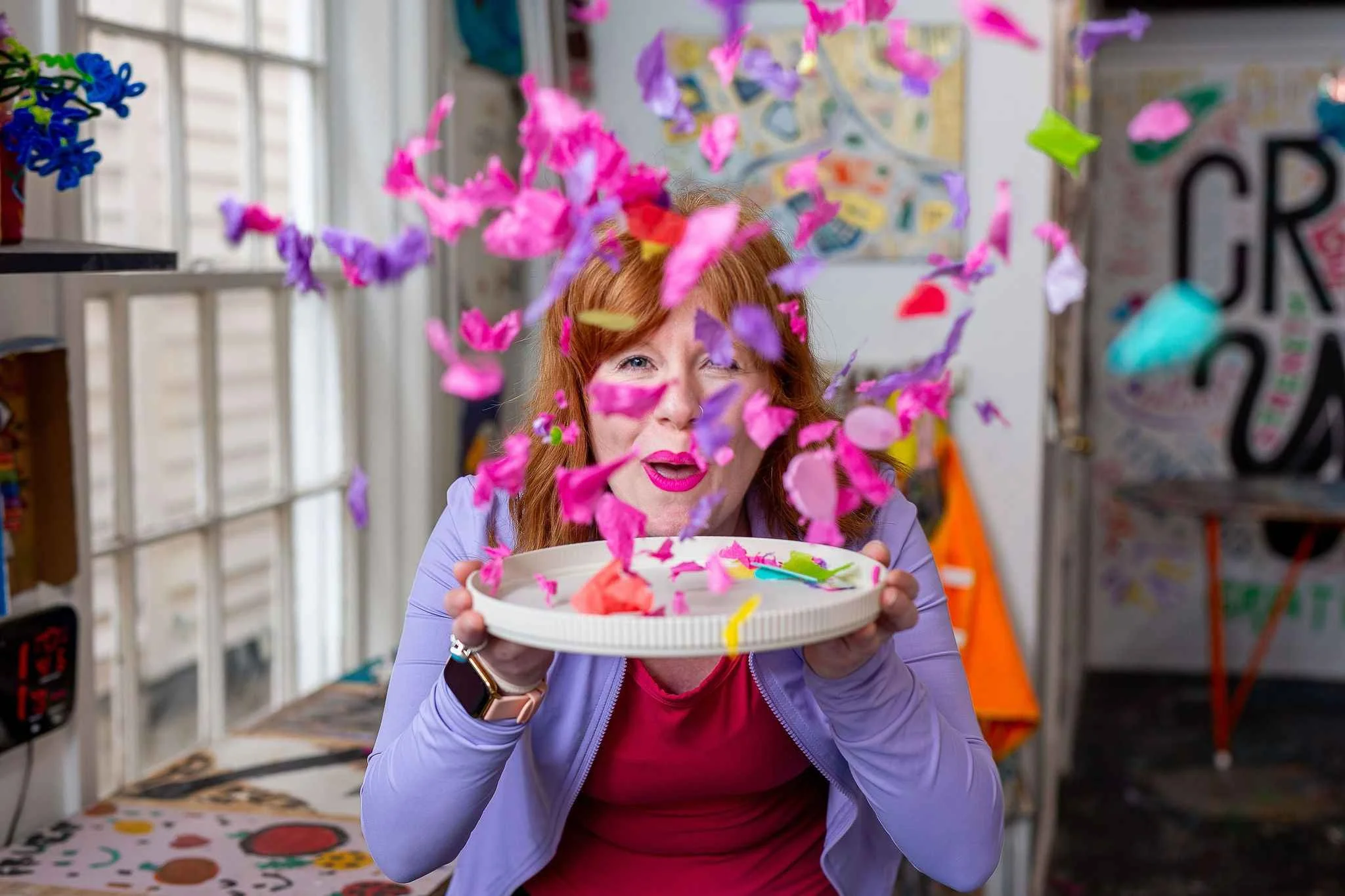 A woman with red hair and pink lipstick is holding a bowl of colorful paper confetti and smiling. The confetti is mid-air, falling around her, creating a festive atmosphere. The background shows a room with various colorful decorations and art.