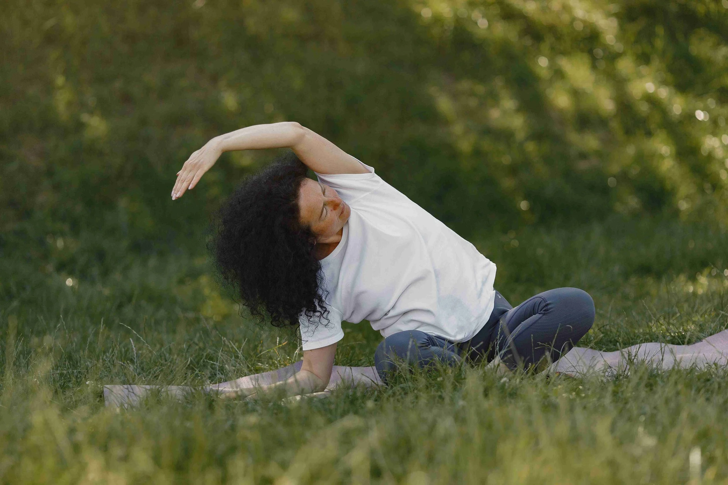 Woman practicing yoga outdoors on grass with trees in the background, performing a side stretch while sitting on a yoga mat.