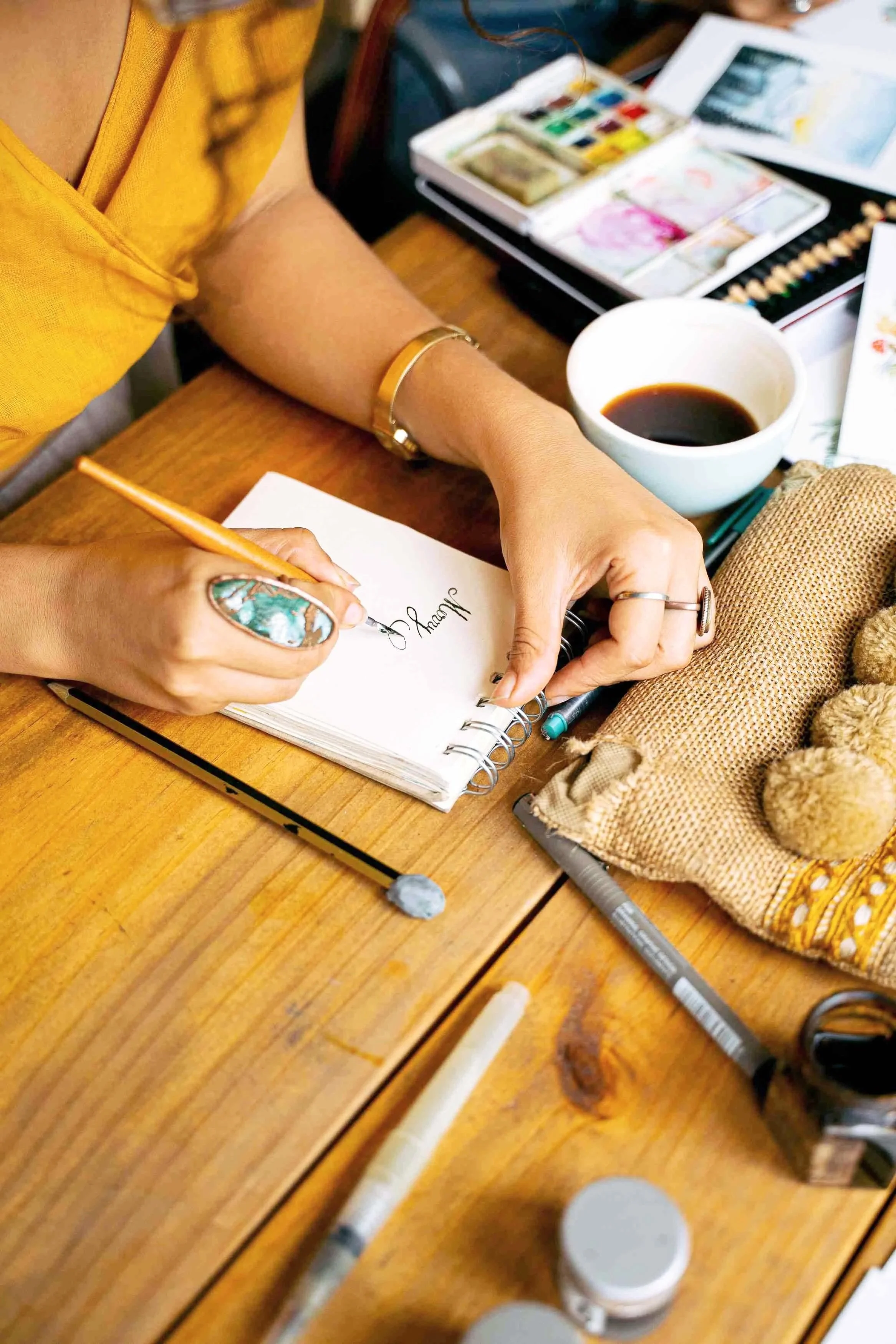 Close-up of a person's hand writing in a notebook, surrounded by art supplies including watercolor paints, colored pencils, and a cup of coffee, on a wooden table.