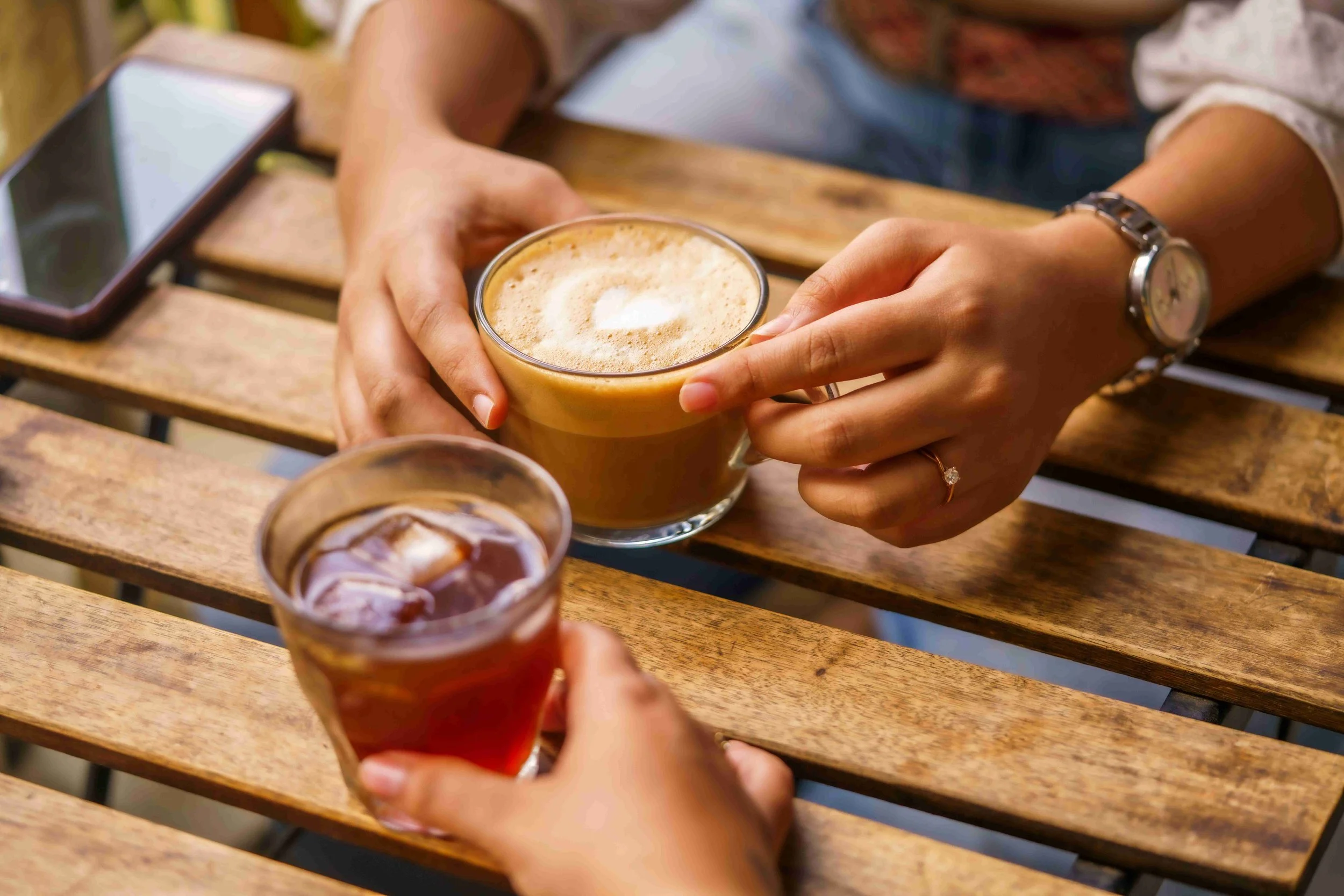Two people holding coffee cups, one with a latte and the other with a tea, sitting at a wooden table