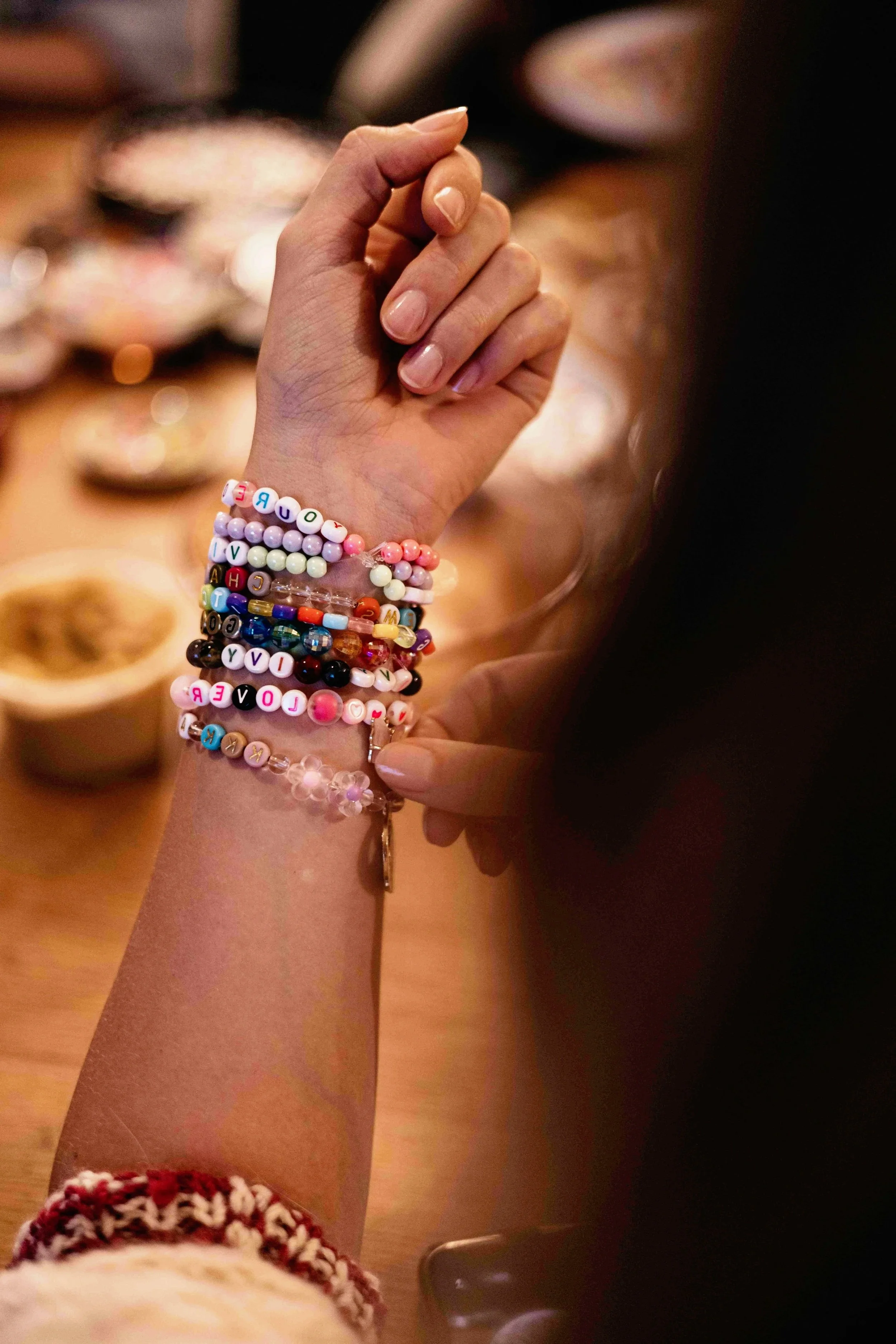 A person's hand and wrist are visible, adorned with multiple colorful beaded bracelets with letters spelling out words, including some that spell "LOVE" and other positive messages.