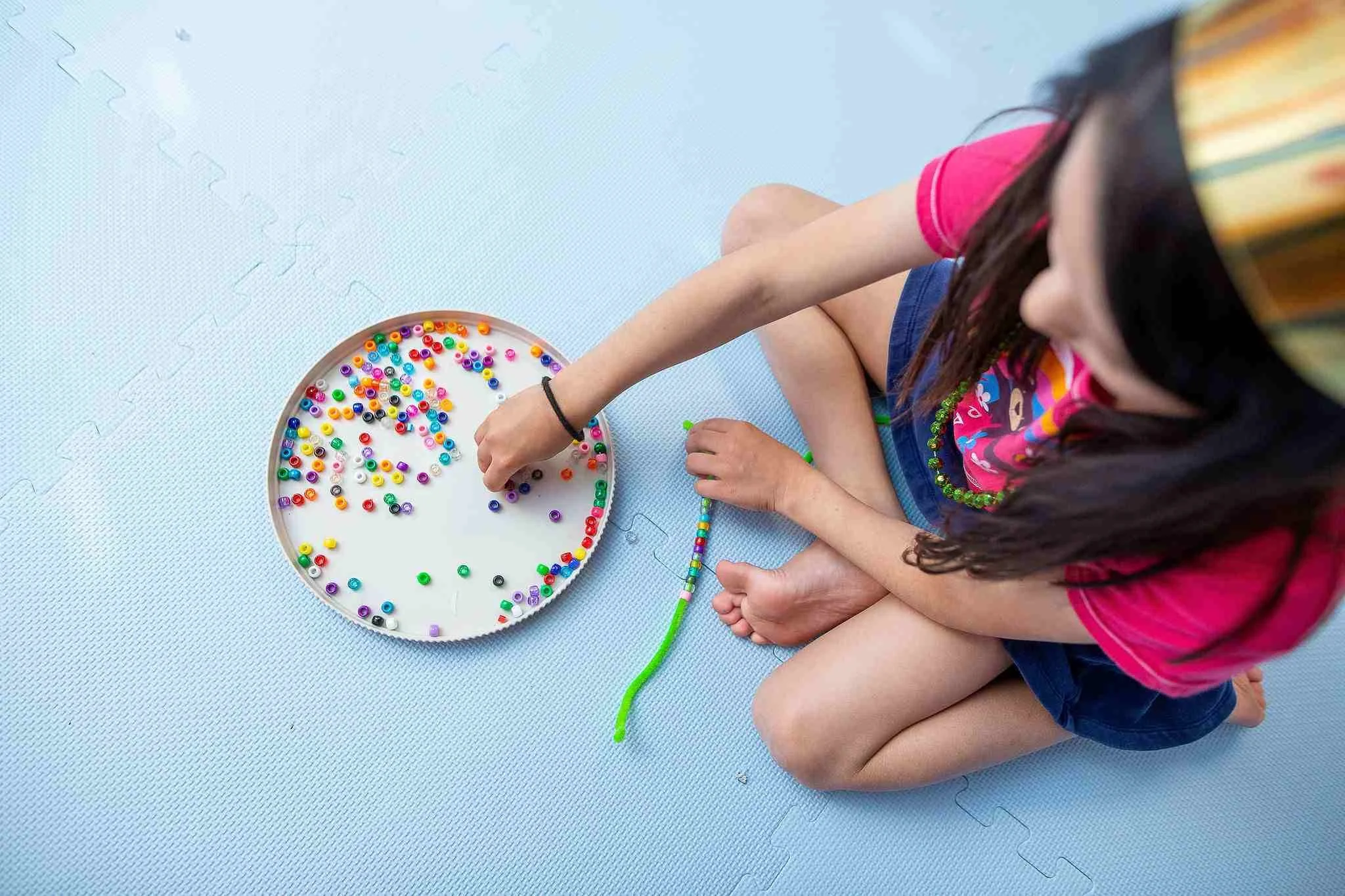 A young girl sitting on a blue mat, reaching for colorful beads on a white circular tray, with some beads strung into a bracelet in front of her.