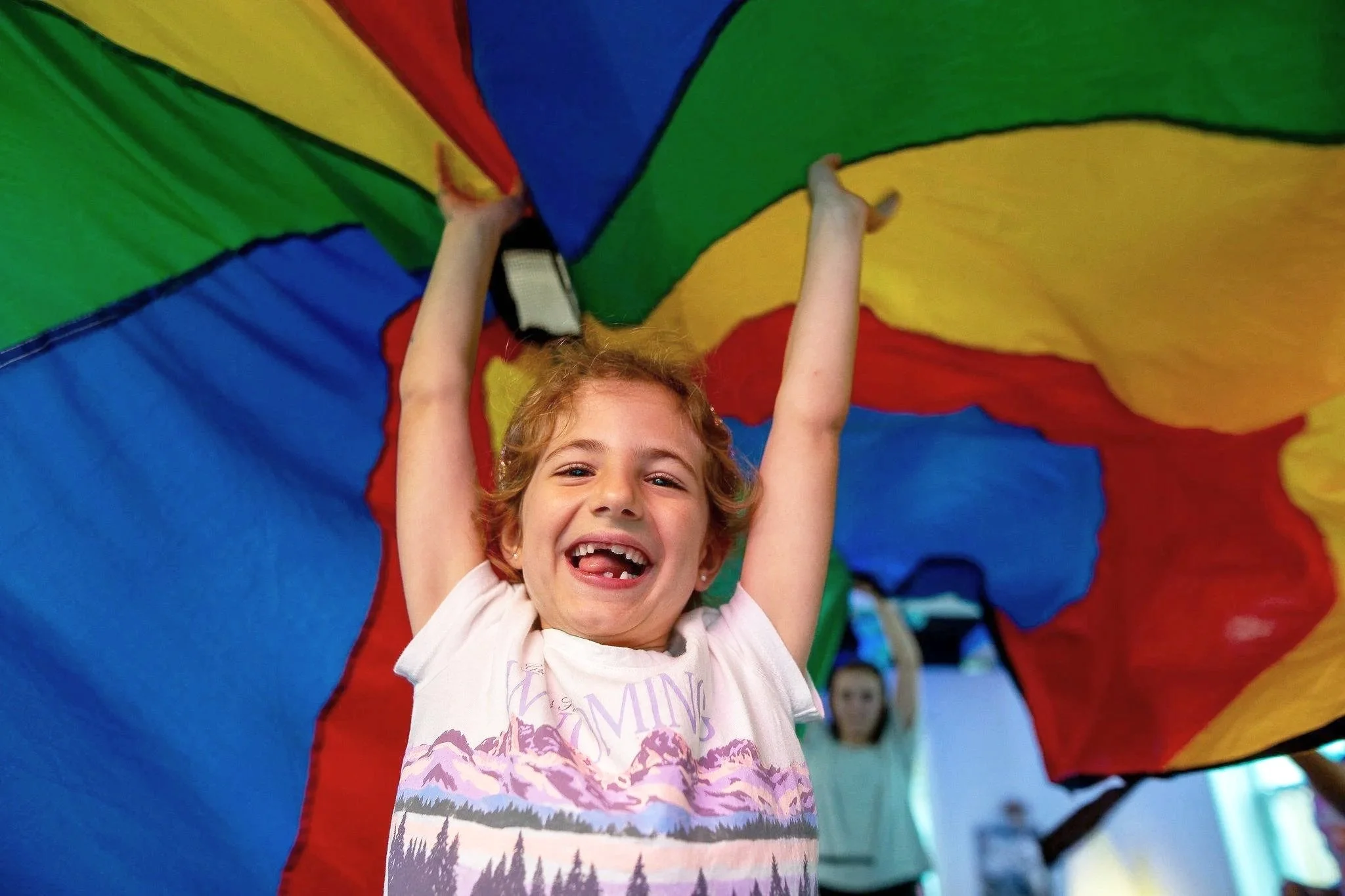 A young girl with curly red hair smiling and lifting her arms, standing under a colorful parachute. Other children are visible in the background.