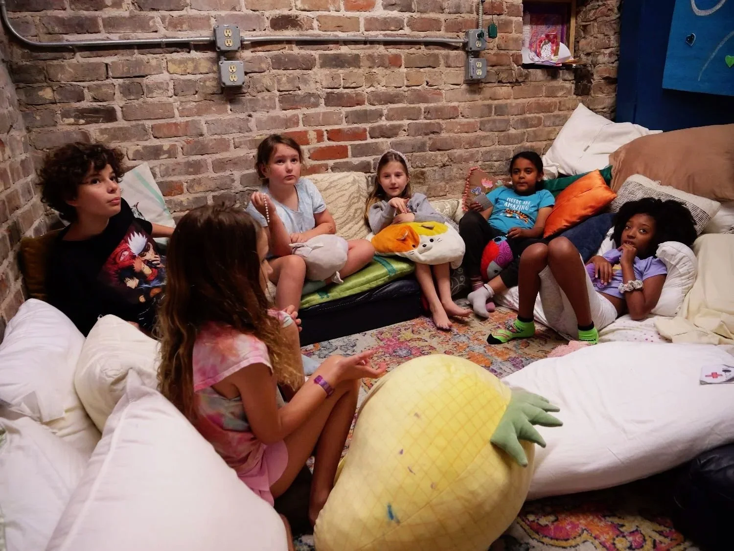 Group of children sitting on couches and cushions in a cozy room with exposed brick wall, talking to each other.