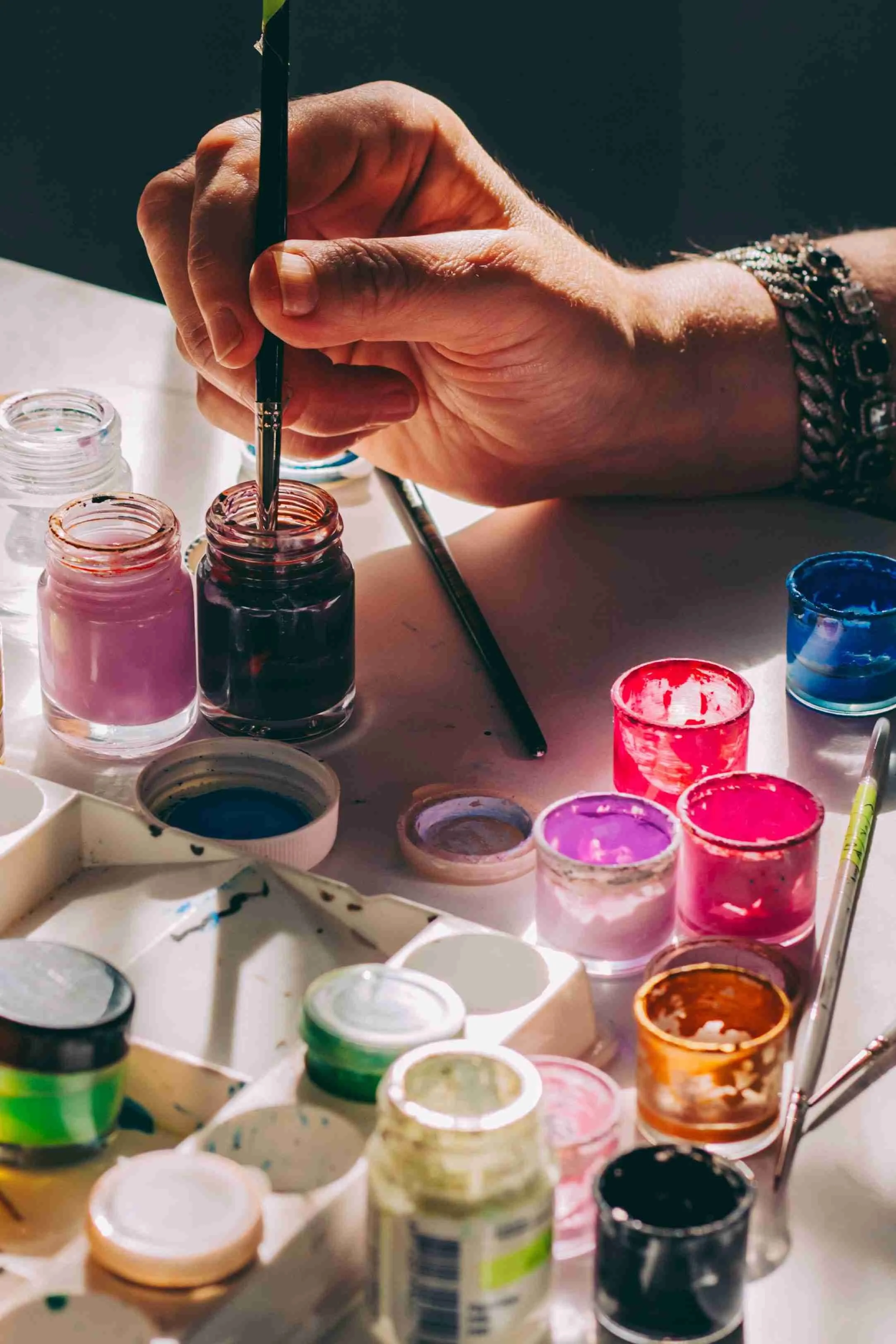 A person's hand holding a small paintbrush over jars of pink, black, and red paint, surrounded by various opened paint containers on a white surface.
