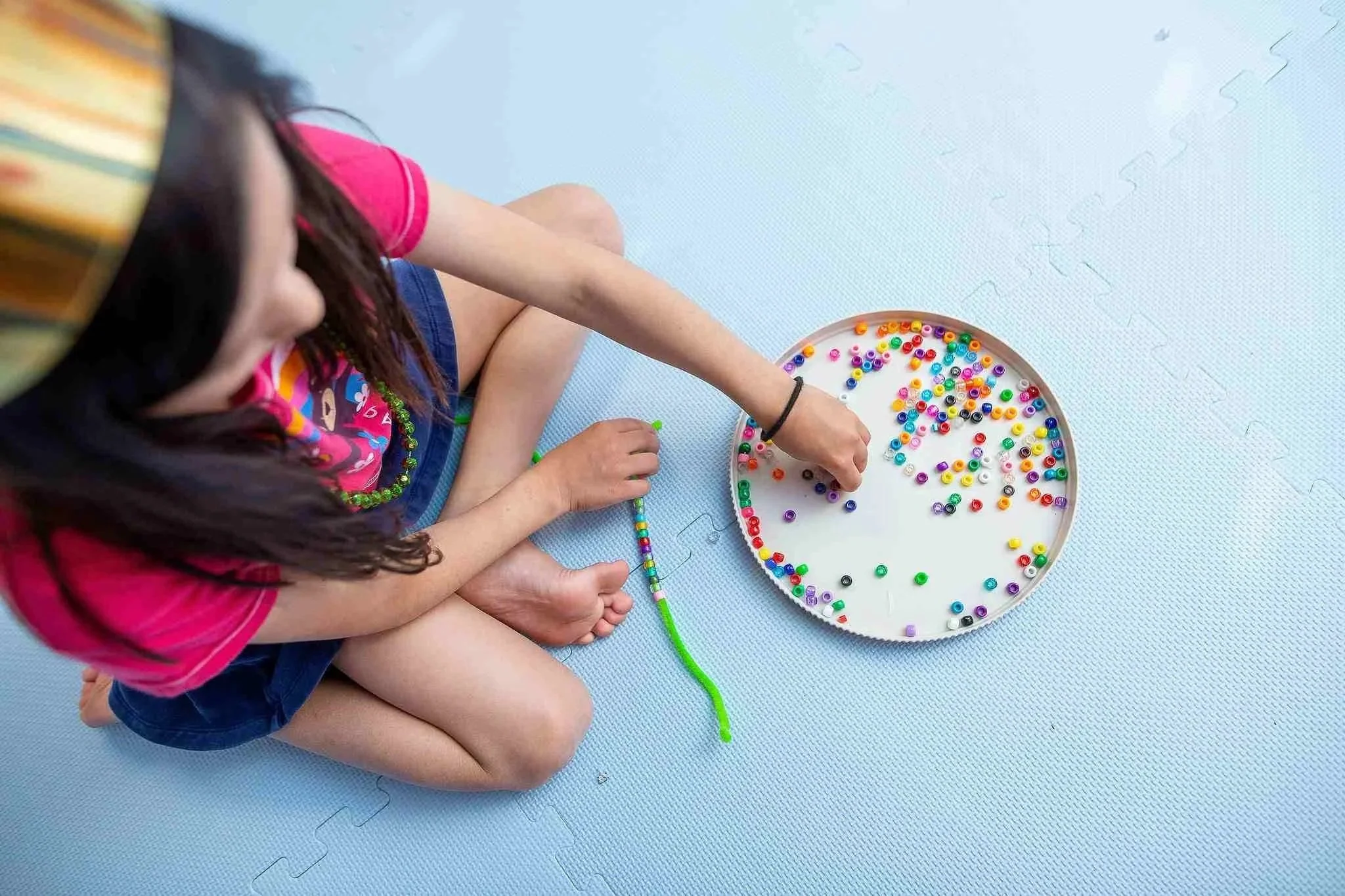 A young person sitting on the floor, wearing a pink shirt and blue shorts, engaging in bead crafting activity. They have a circular tray filled with colorful beads and are stringing beads onto a green string stretched across their lap. The surface is a light blue foam mat.