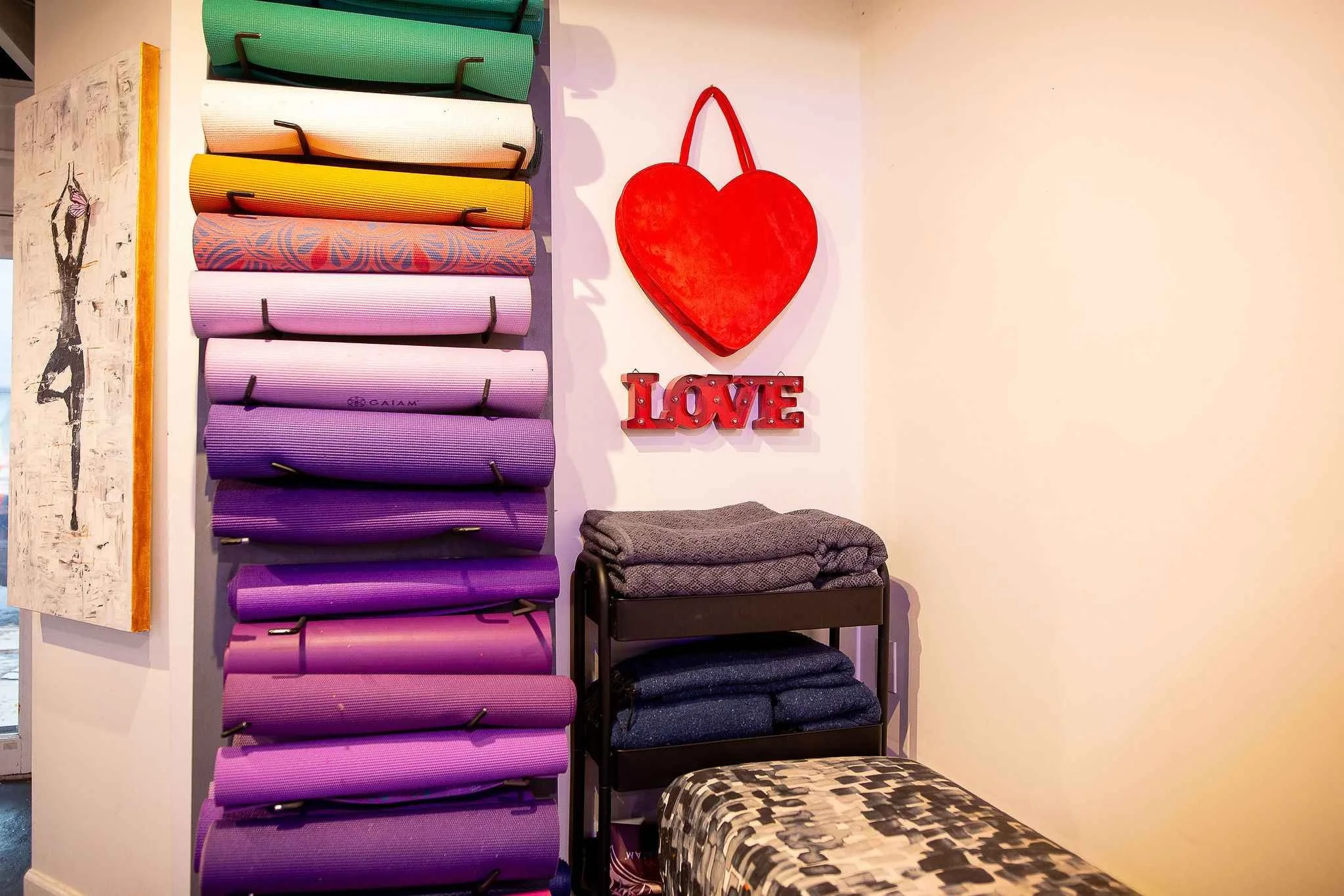 Display of colorful yoga mats on shelves next to a wall with a red heart decoration and a sign that says 'LOVE' in lights. There are folded yoga blankets and bolsters on a black table below the mats.